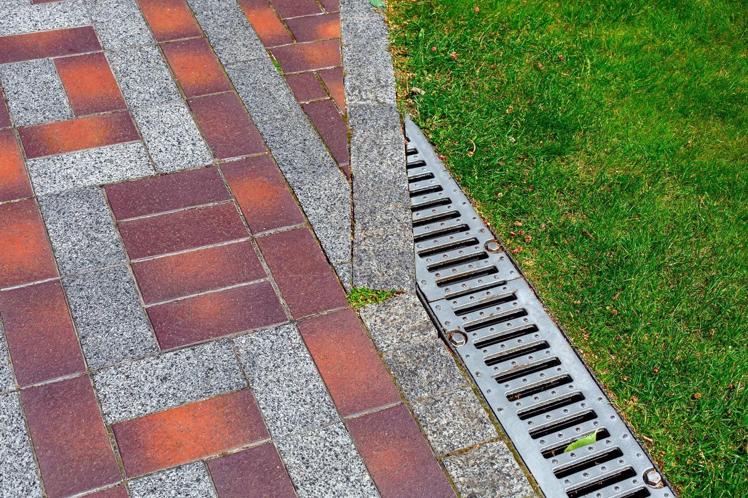 Brick walkway beside a metal storm drain grate bordering green grass