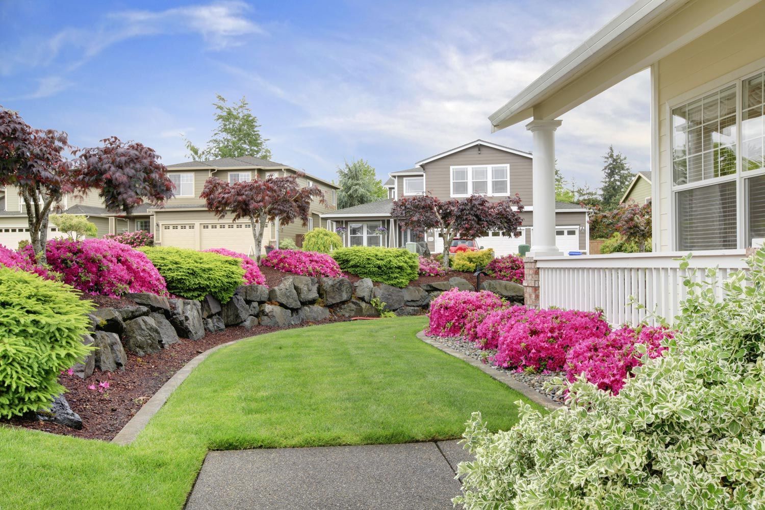 Colorful landscaped garden with pink flowers, green lawn, stone path, and a white house porch