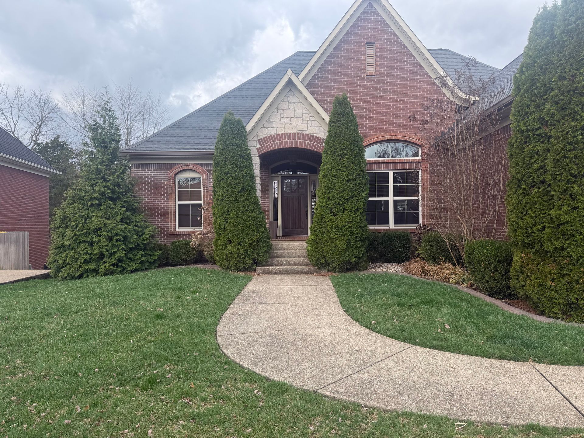 Brick house with a curved concrete walkway, manicured lawn, and tall evergreen shrubs by the entrance