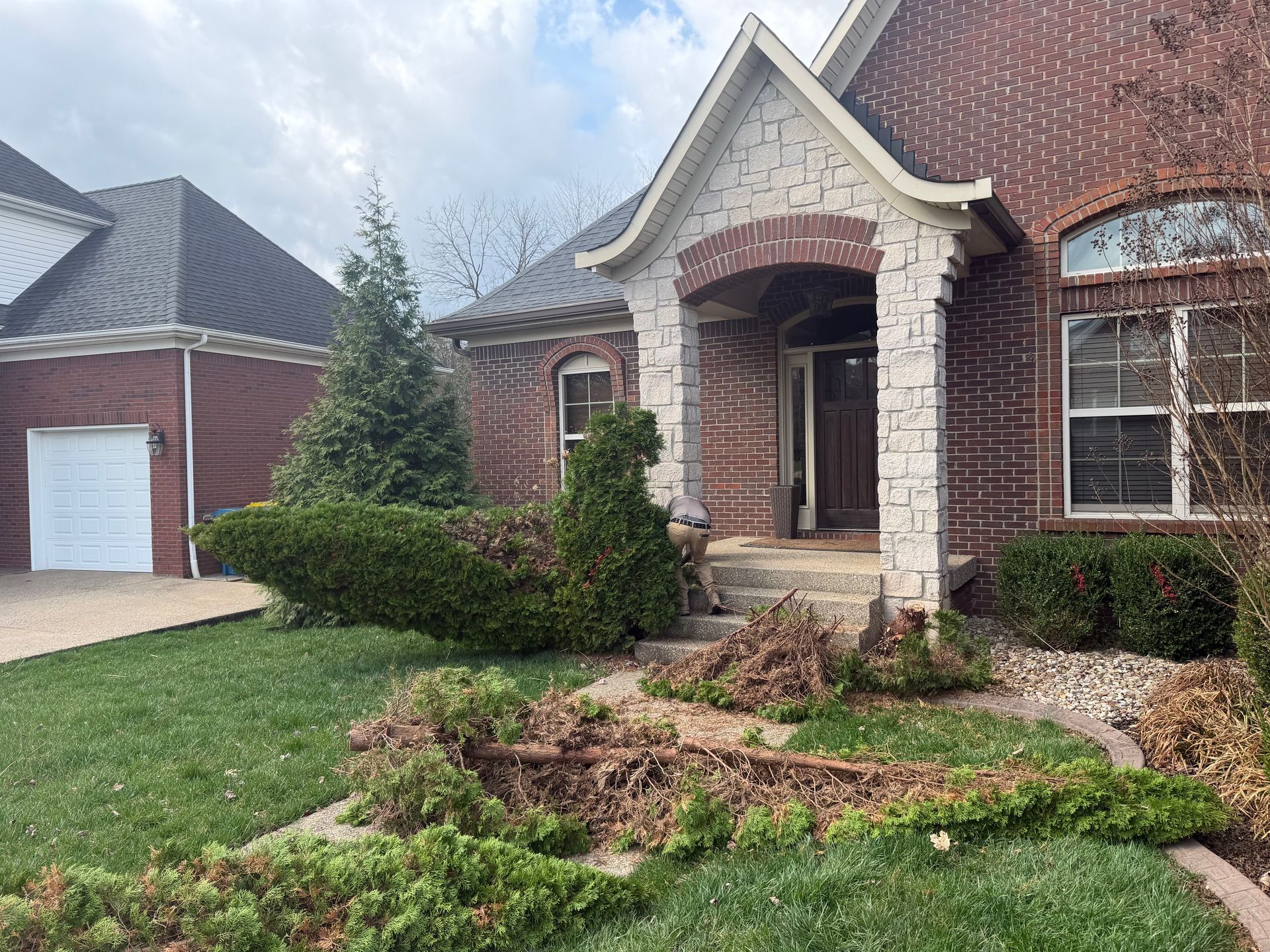 Brick house with a front porch, arched entry, garage, and landscaped yard with a fallen tree branch.