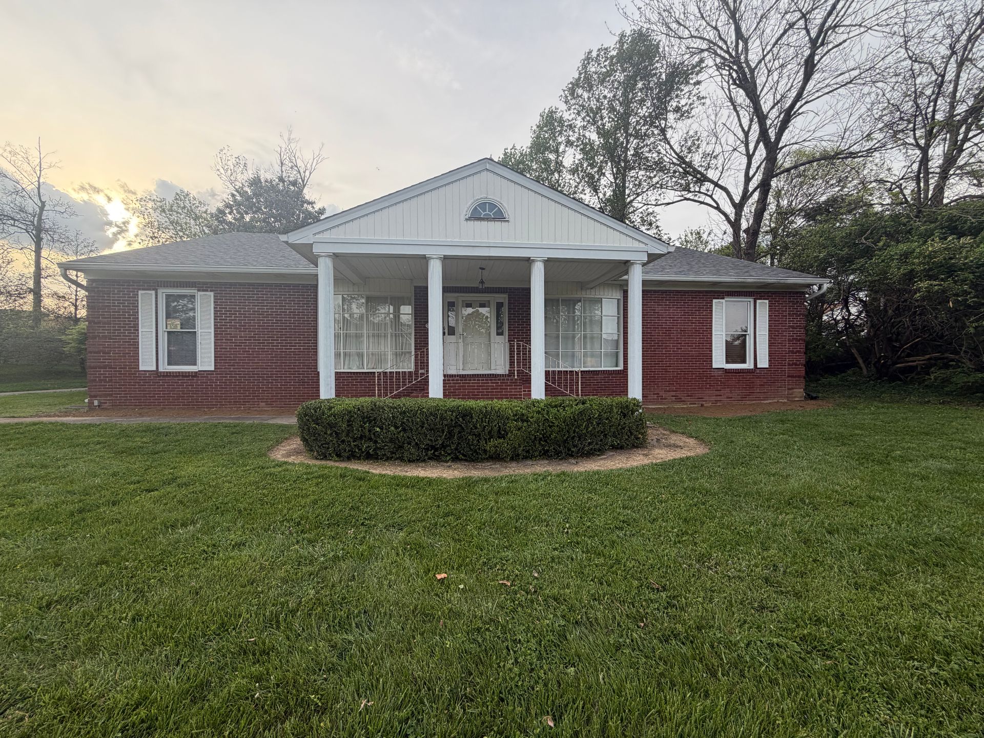 Small red brick house with white columns and a front porch, surrounded by a green lawn at dusk