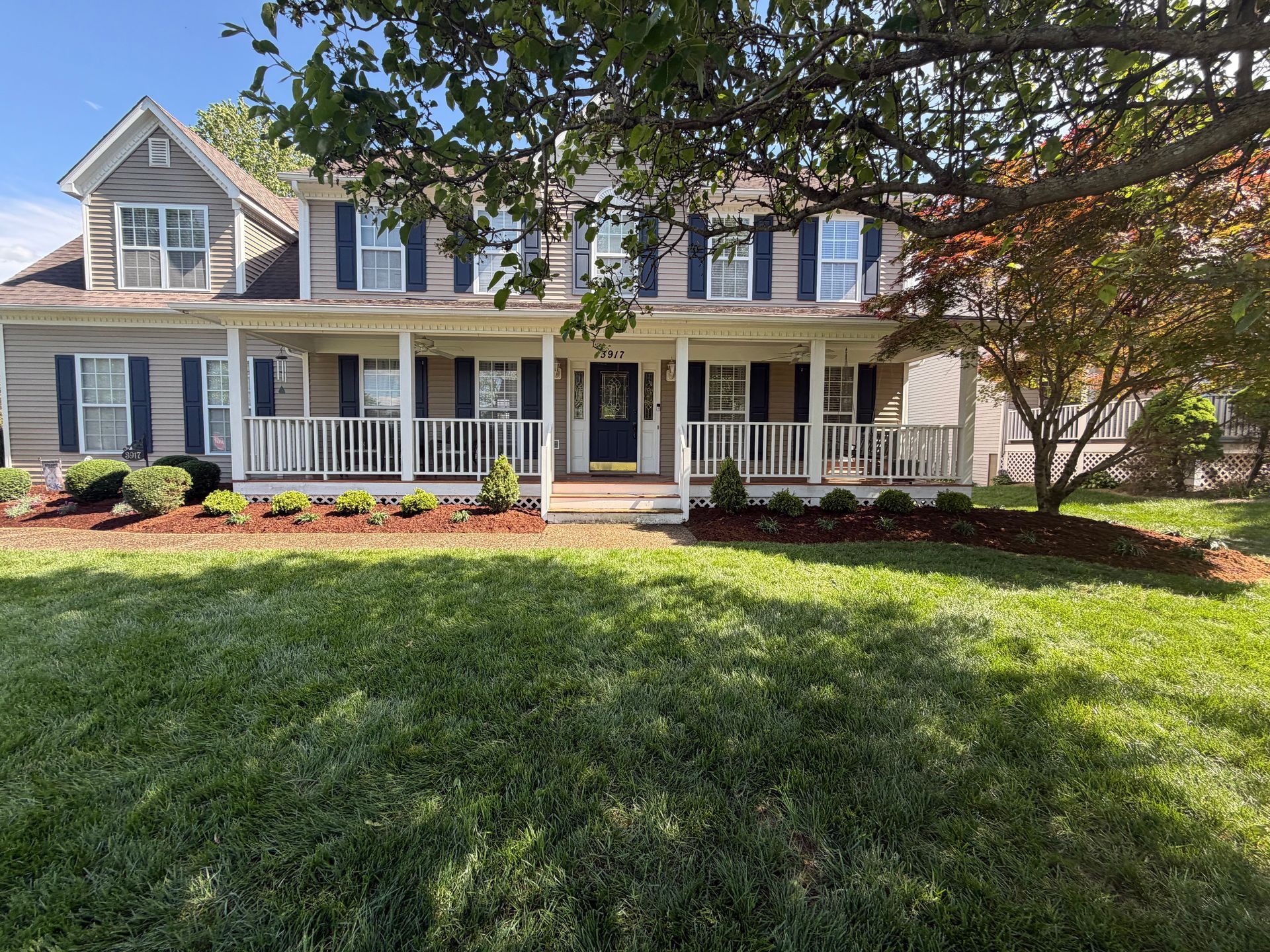 Two-story blue-gray house with white porch, shutters, and a green front lawn under trees