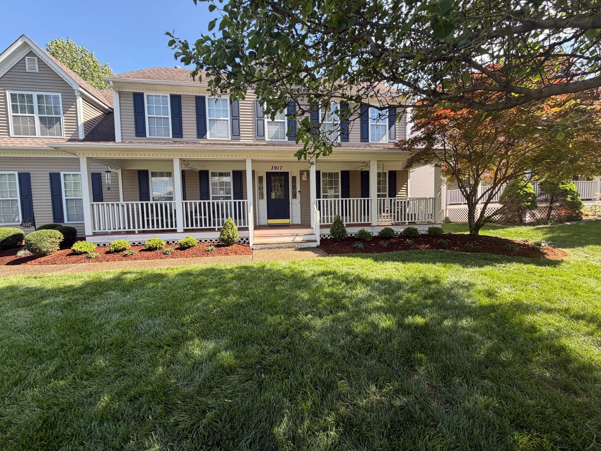 Two-story suburban house with blue shutters, white porch, and green lawn under trees