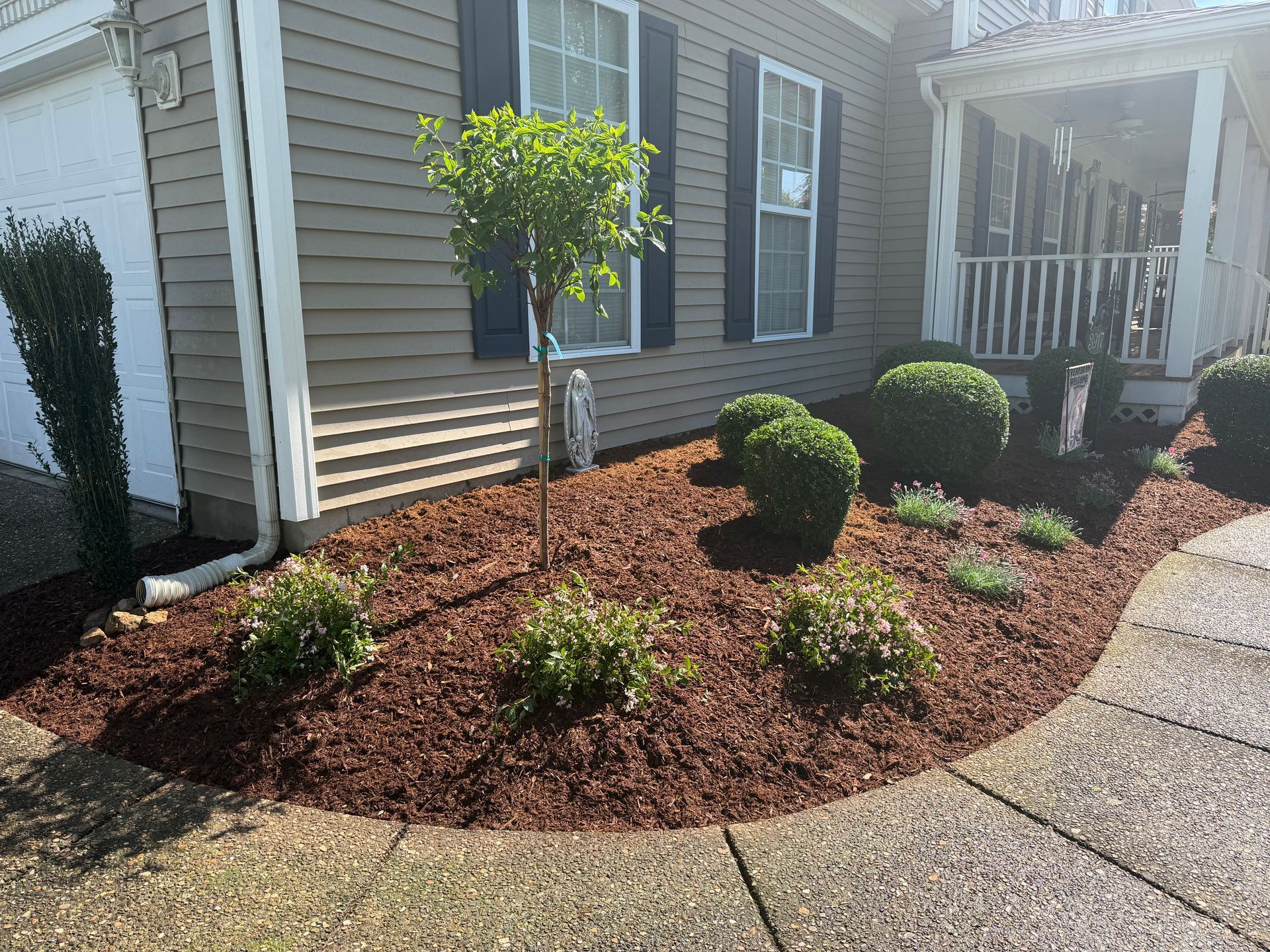 Front yard landscaping with mulch beds, small shrubs, a young tree, and a house porch beside a curved driveway