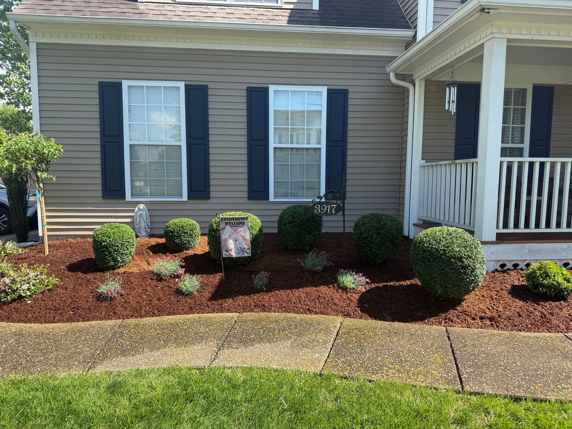 Front yard of a house with blue shutters, porch, trimmed shrubs, and a mulched flower bed along the sidewalk.