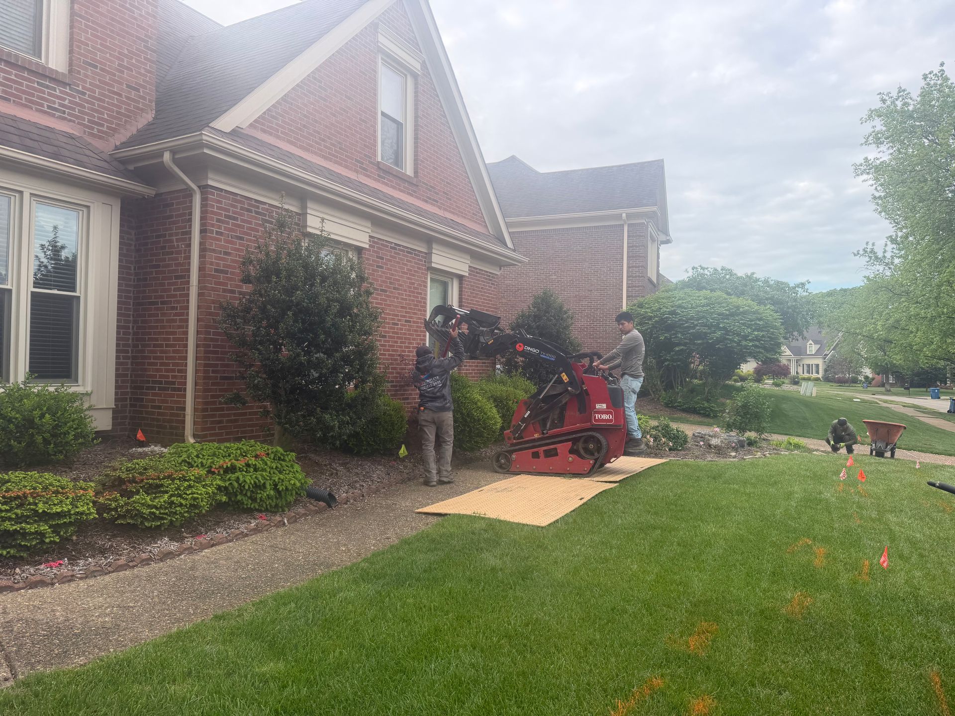 Person working on a red lawnmower beside a brick house with a green lawn and garden path