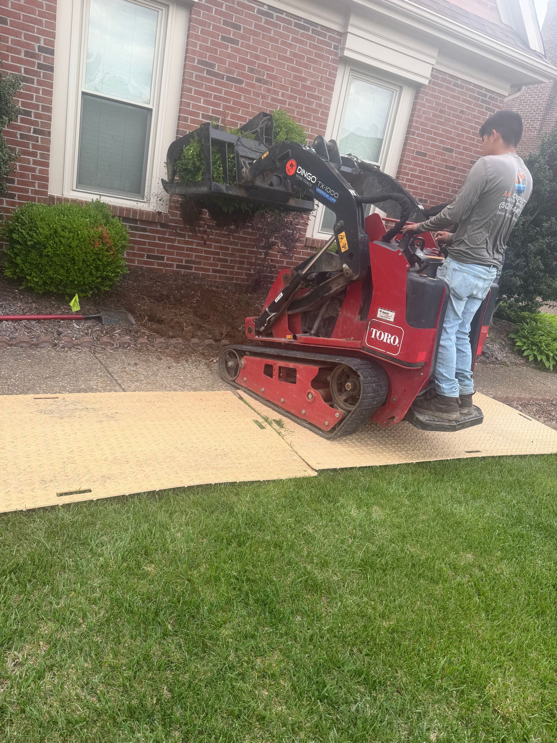 Worker operating a red Bobcat loader beside a brick house with wet sidewalks and lawn.