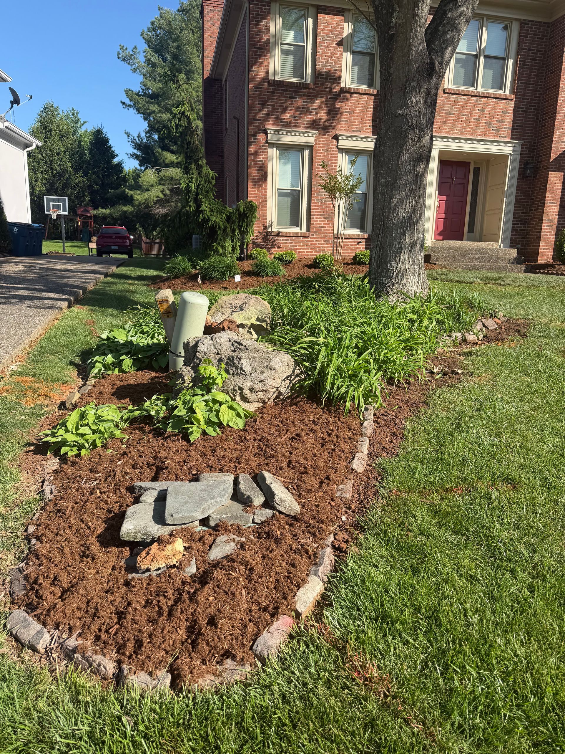 Flower bed with fresh mulch, rocks, and green plants beside a brick house and lawn.
