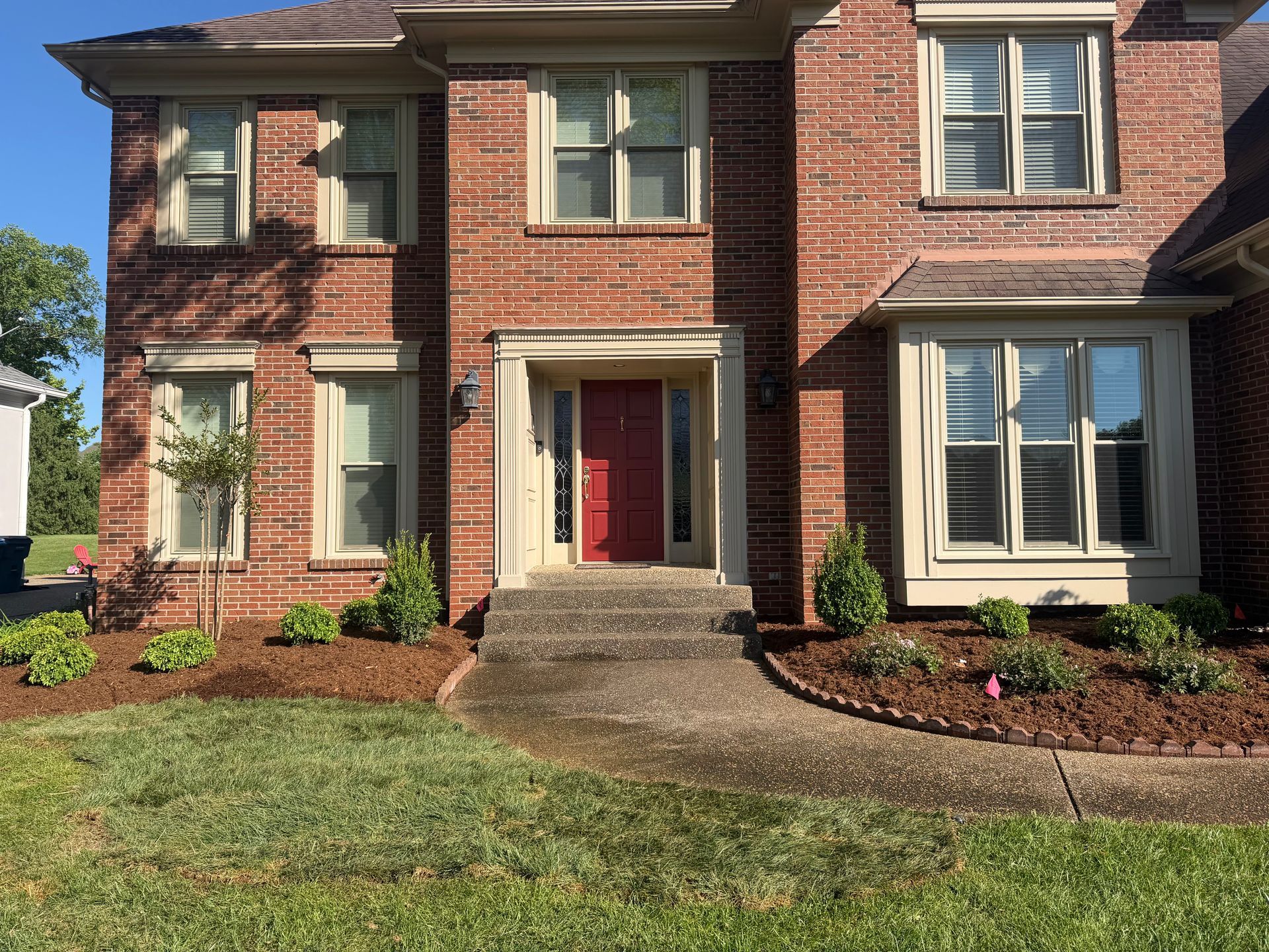 Two-story brick house with a red front door, white columns, and a neatly landscaped front lawn.