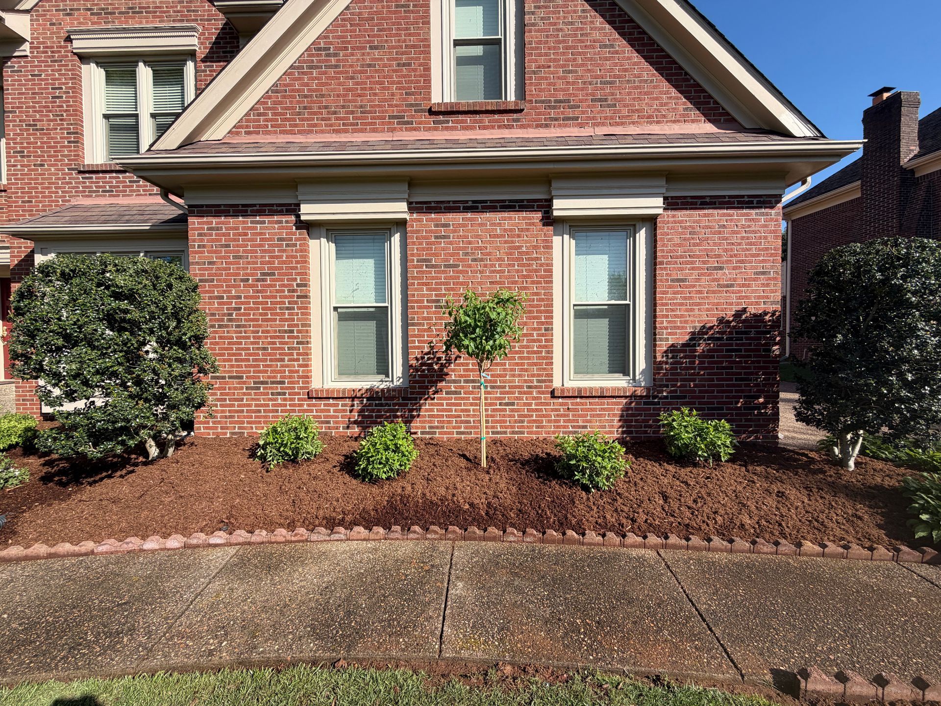 Brick house front with windows, shrubs, and a sidewalk in front