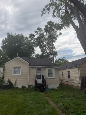 Small beige house with a front porch and overgrown yard under a cloudy sky