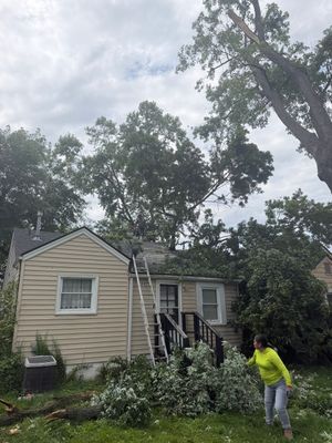 Storm damage in a yard, with fallen tree branches beside a beige house and a person in a bright yellow jacket.