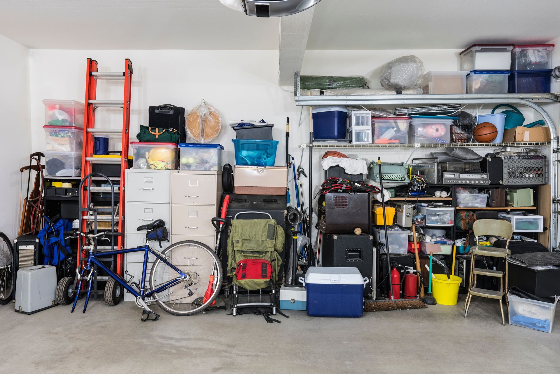 Cluttered garage with stacked storage containers, tools, a ladder, and a bicycle.