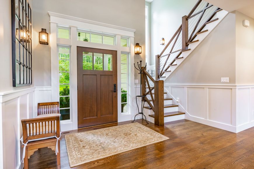 Wooden door and stairs in a brightly lit entry hall with a rug and a chair.