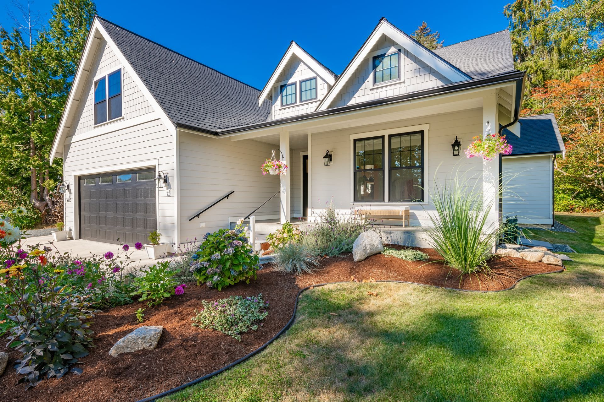 White house with black roof, small front porch, and garage. Landscaped front yard with flowers.