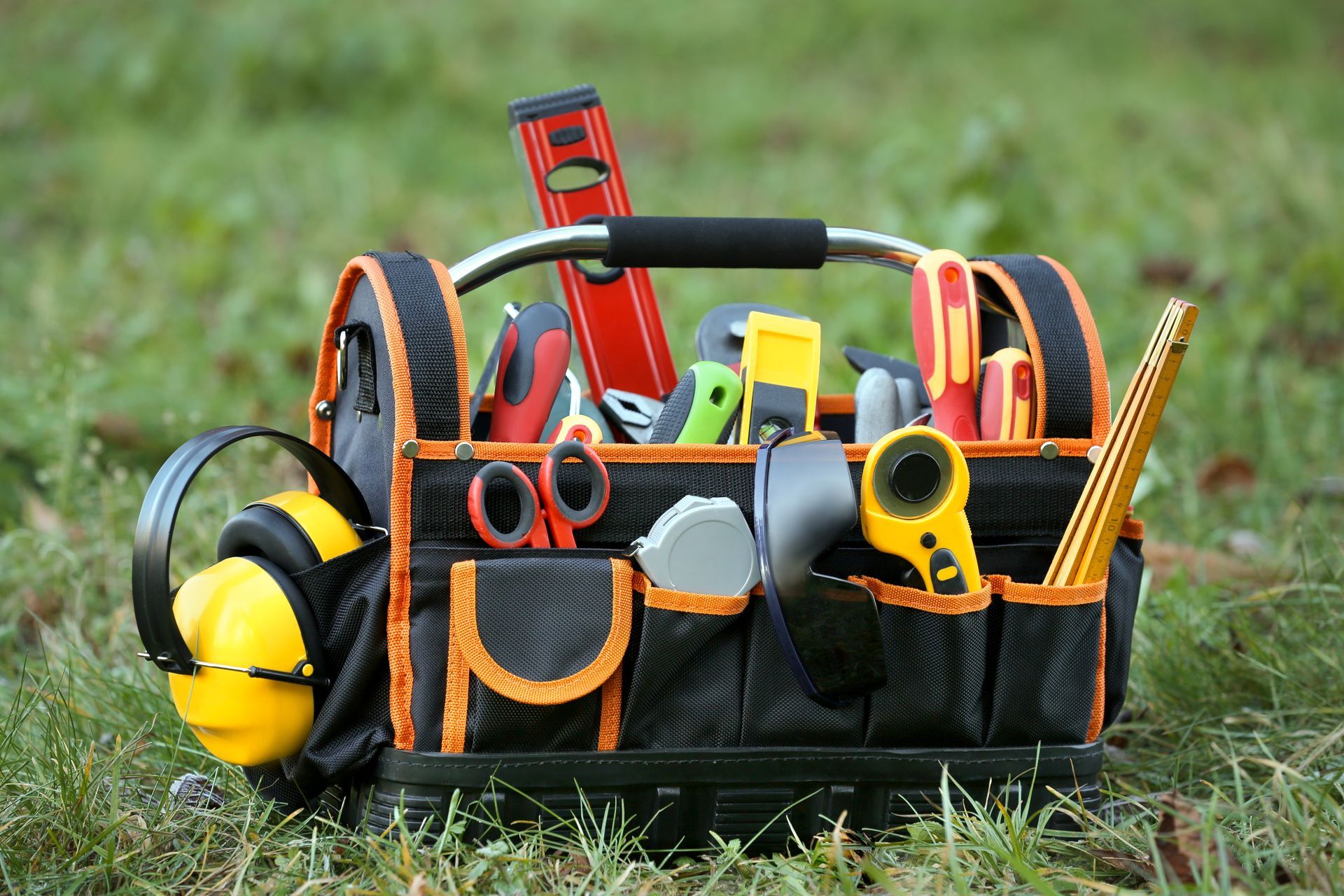Black tool bag on grass, filled with tools like a level, scissors, tape measure, and headphones.