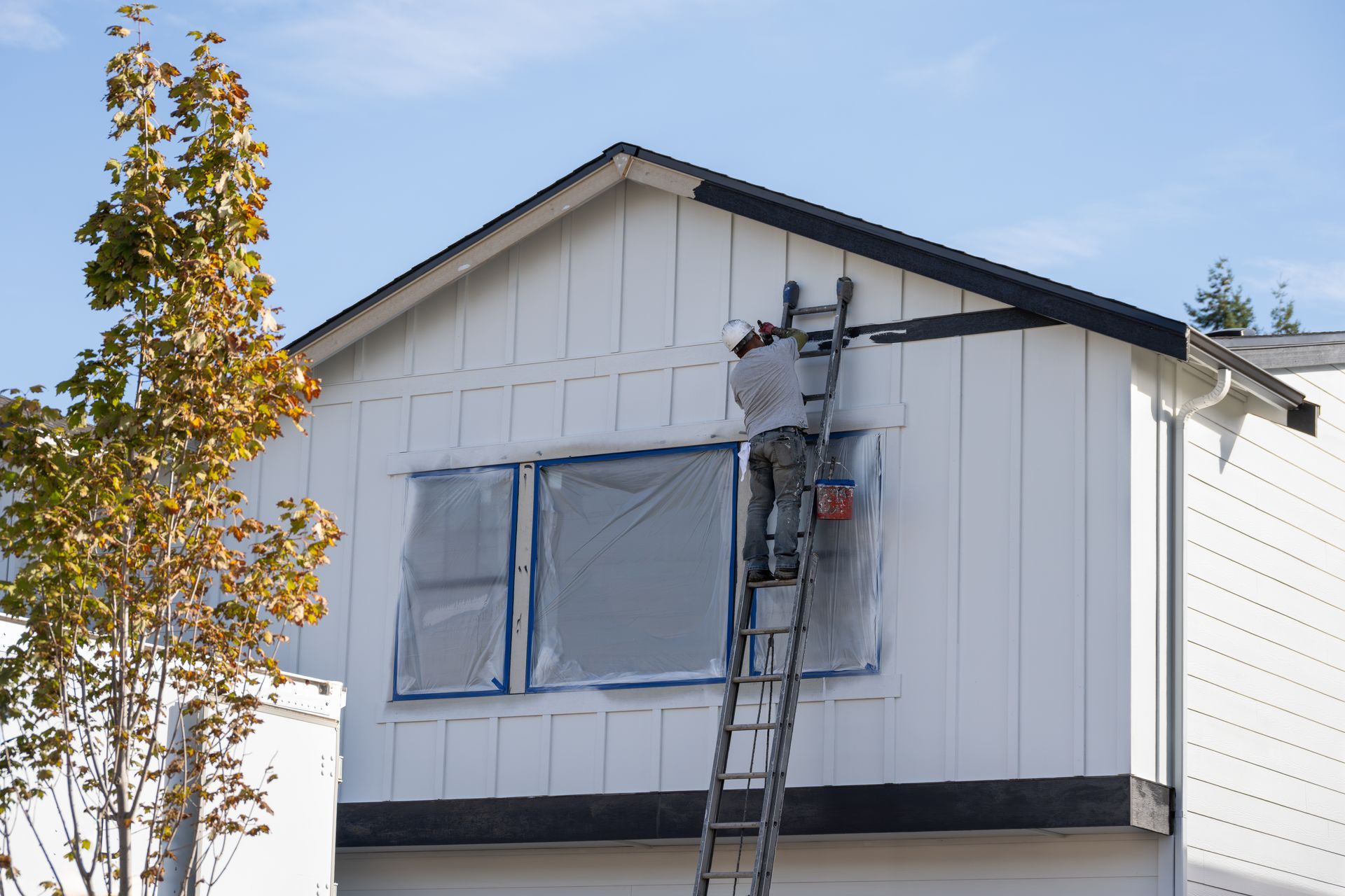Person on ladder painting the white exterior of a house. Windows covered with blue tape. Sunny day.