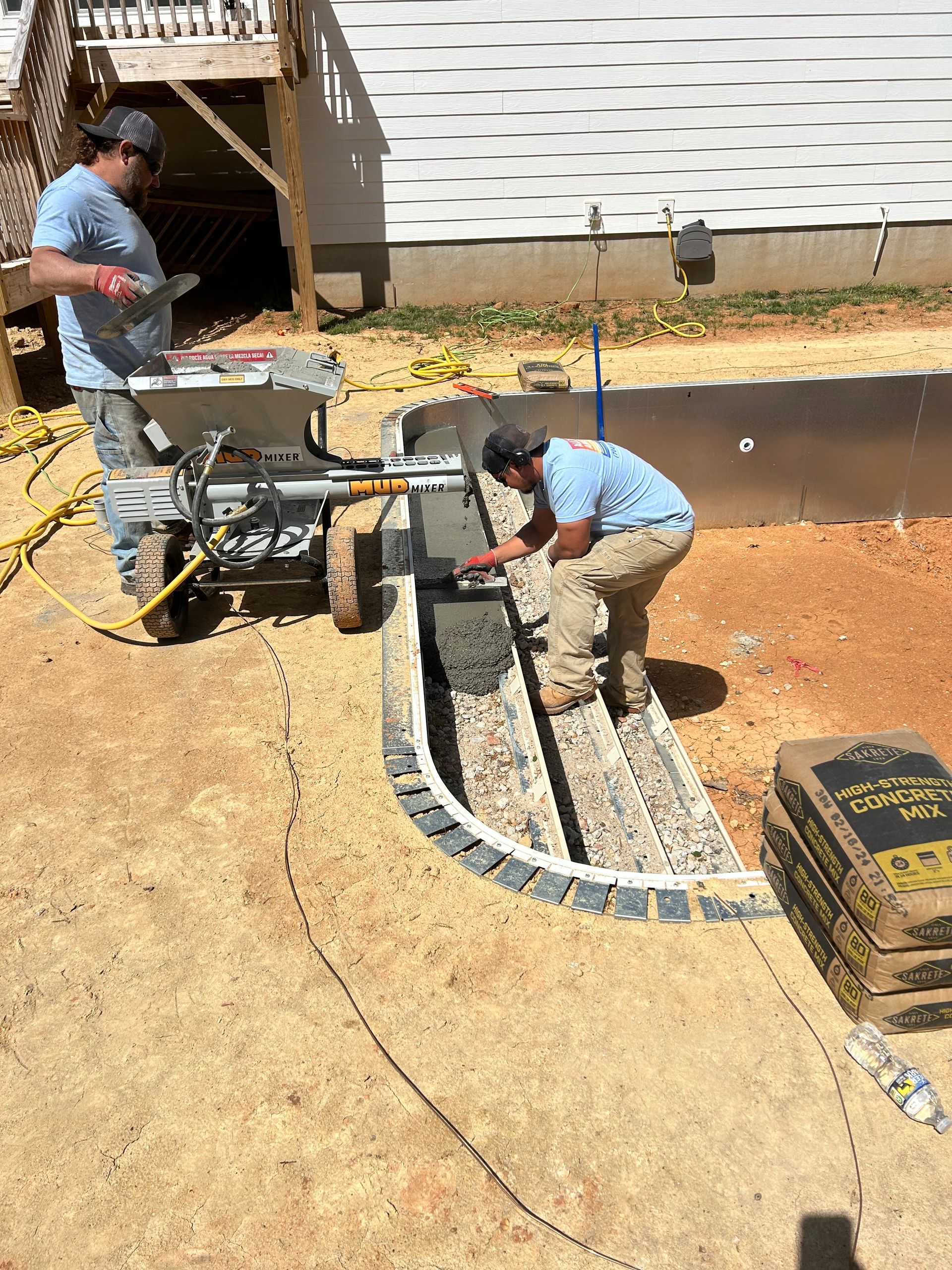 Two construction workers applying concrete to a structure with a power tool, surrounded by cement bags.