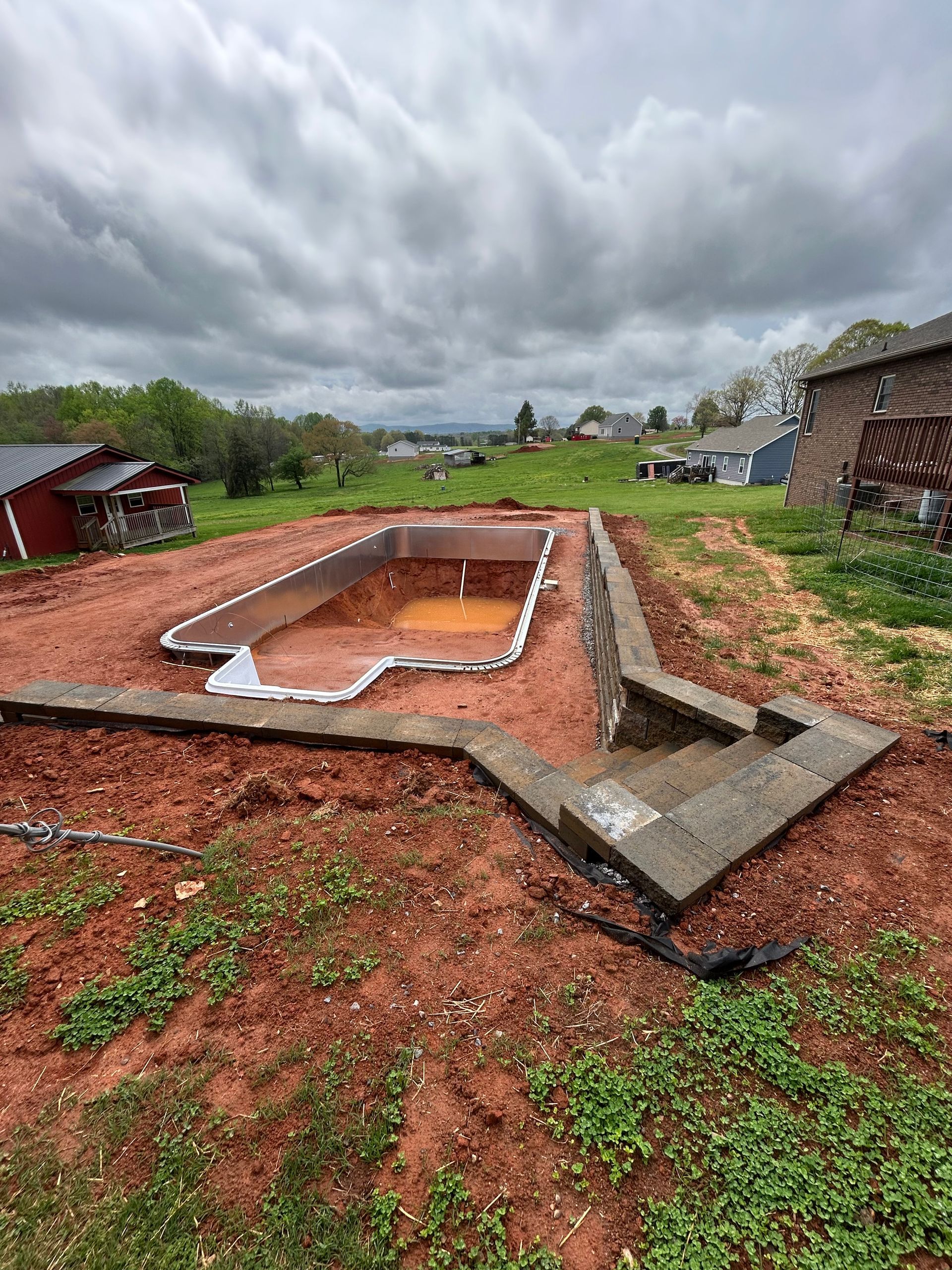 Pool under construction in a yard with red soil, surrounded by concrete and a cloudy sky.