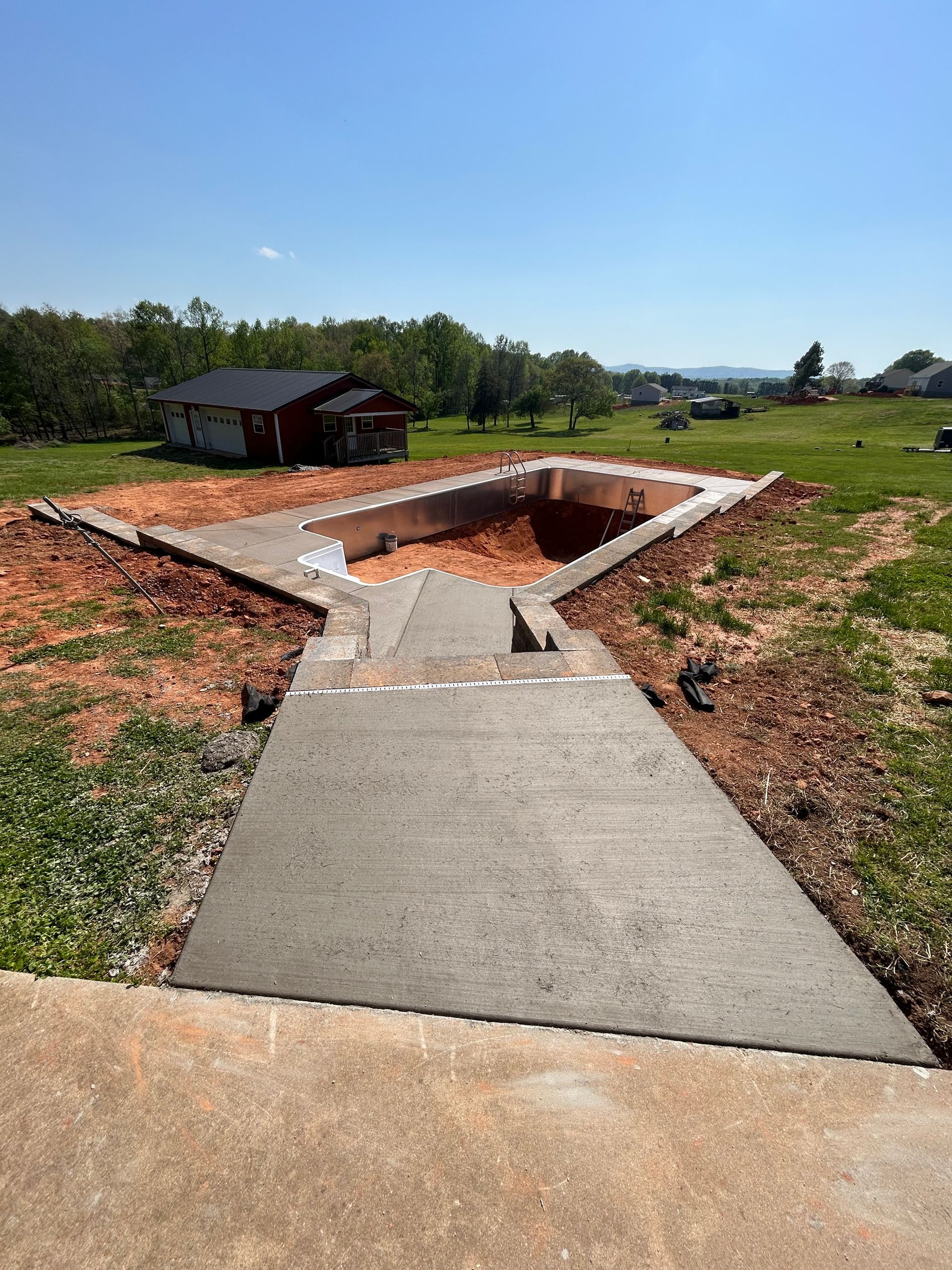 Concrete patio leading to a swimming pool under construction in a grassy area with a house in the background.
