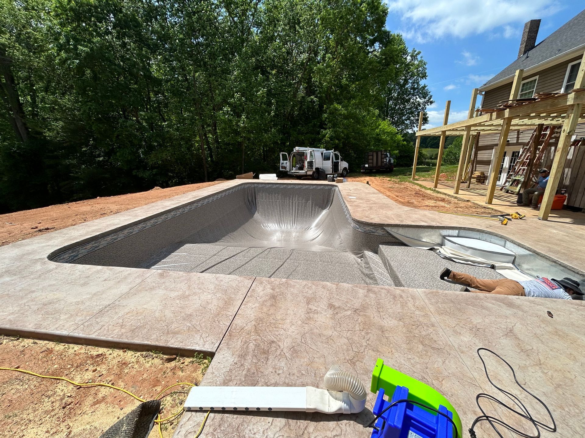 A partially constructed swimming pool with steps, concrete surround, and nearby wood structure; sunny day.