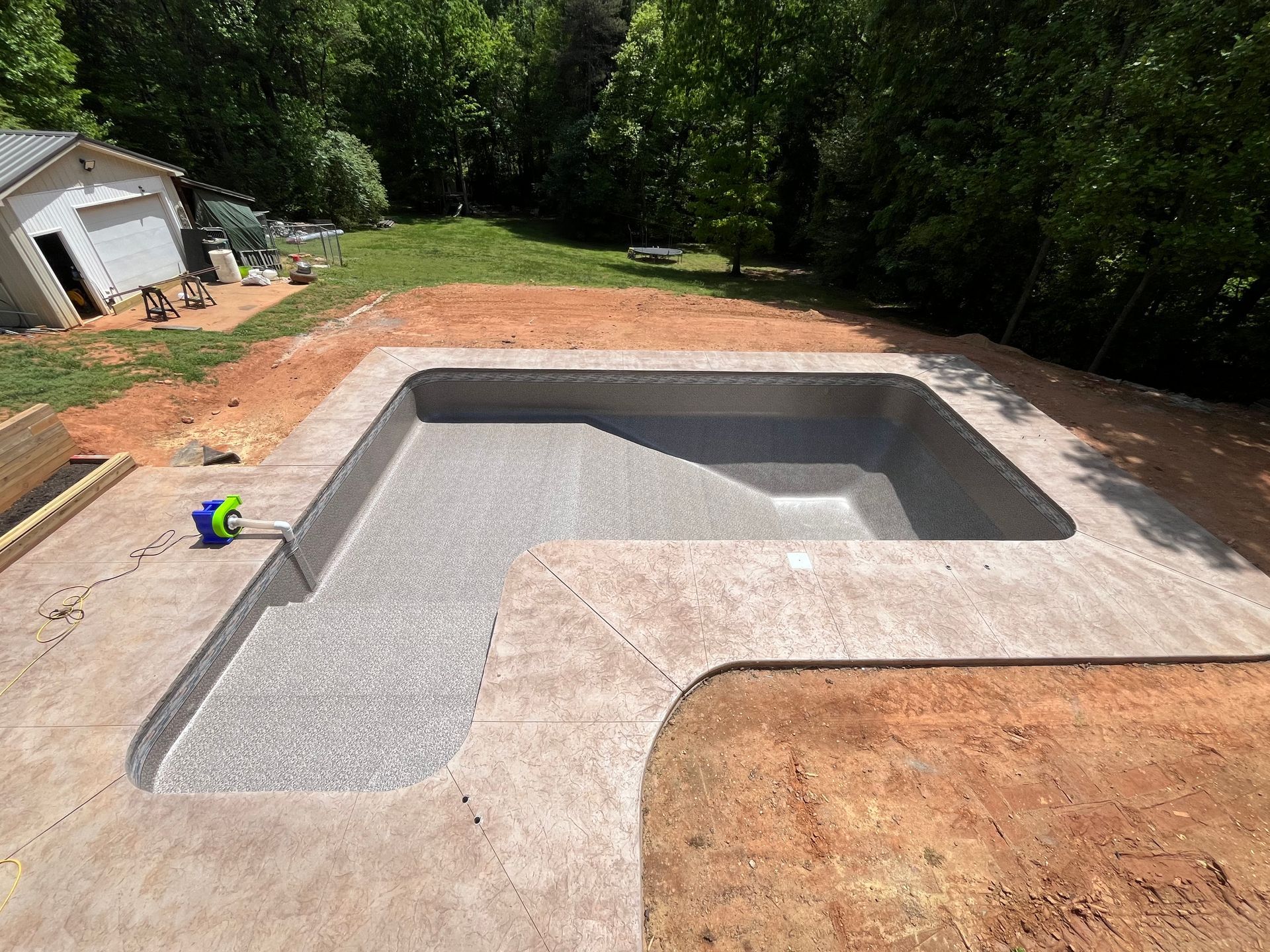 L-shaped concrete pool under construction; gray interior, surrounded by concrete deck and bare earth, trees in background.