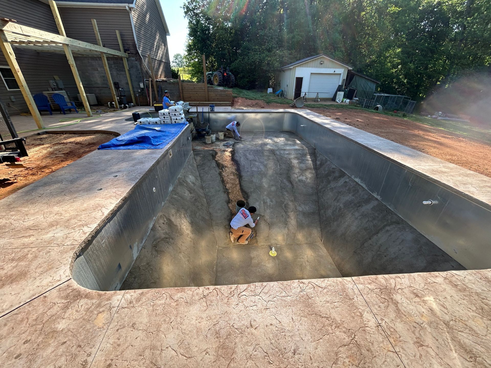 Construction of a pool: workers inside a rectangular pool, surrounded by concrete, with a house and trees in the background.