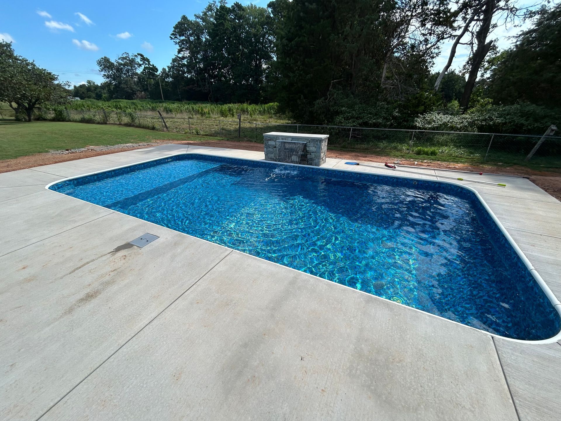 Rectangular pool with blue water, surrounded by concrete. Stone wall feature at one end, trees in the background.
