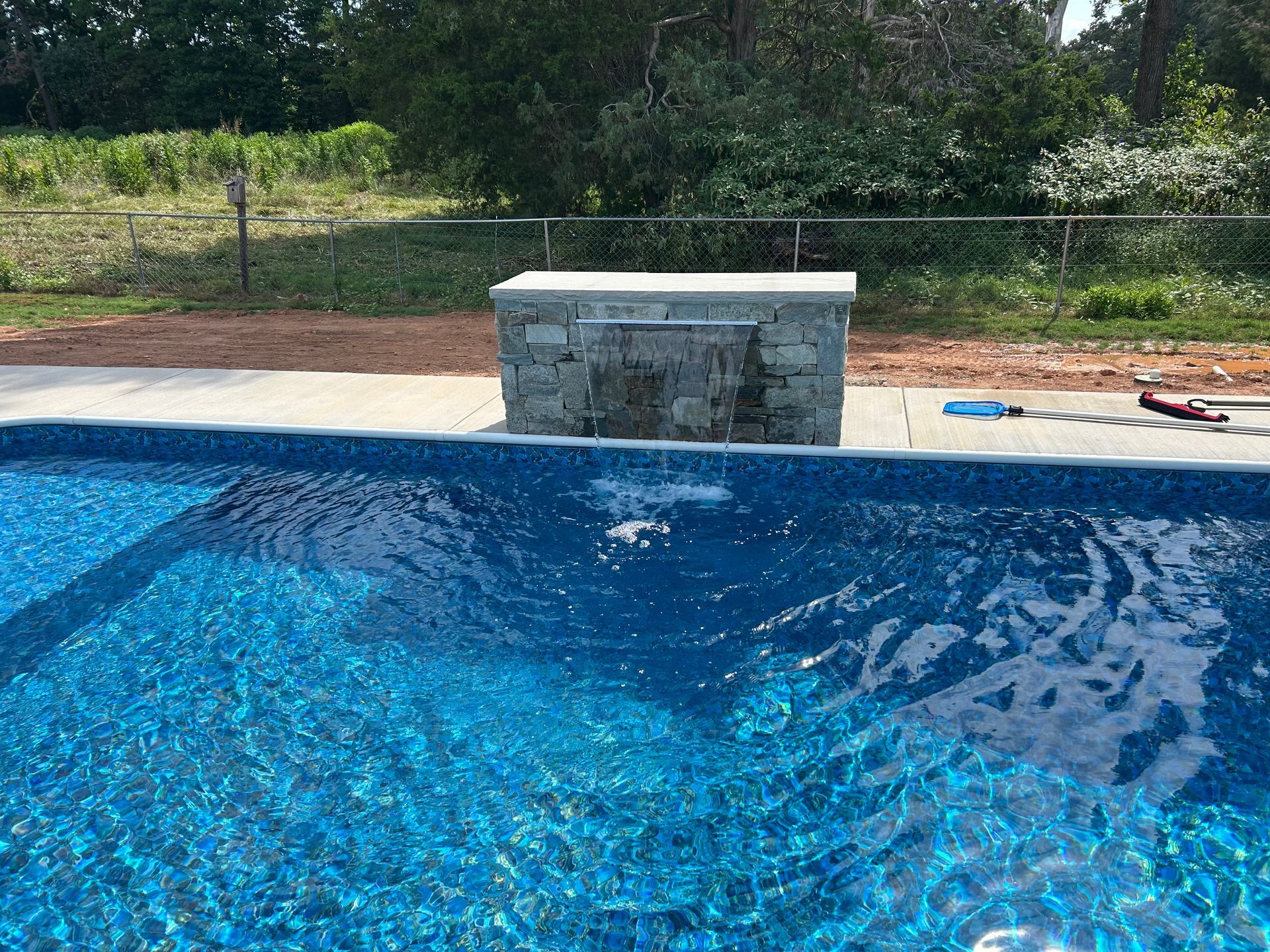 Stone waterfall feature in a blue swimming pool, with a grassy area and trees in the background.