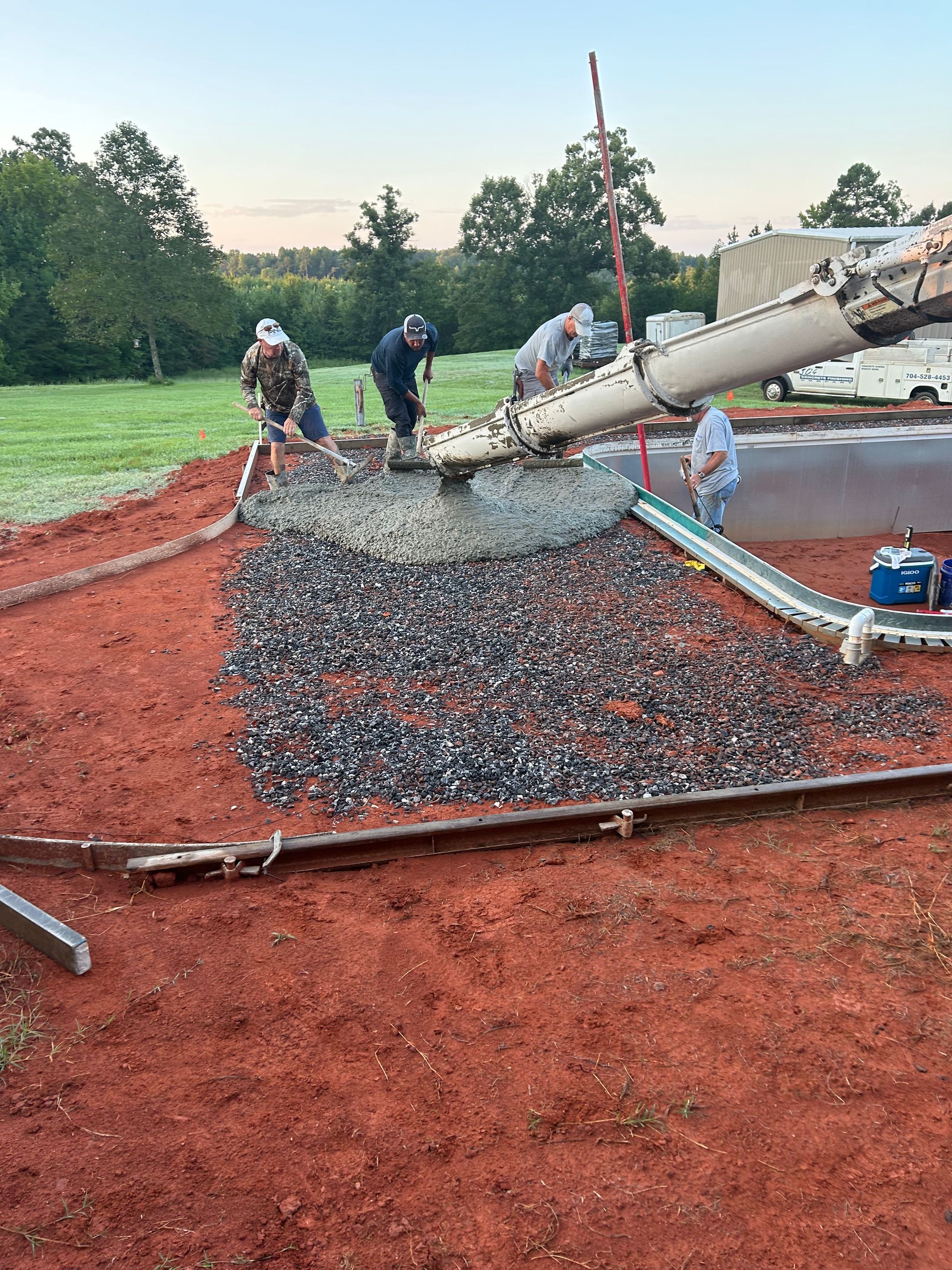 Workers pouring concrete from a truck into a gravel-covered area, red earth surrounds the work area.