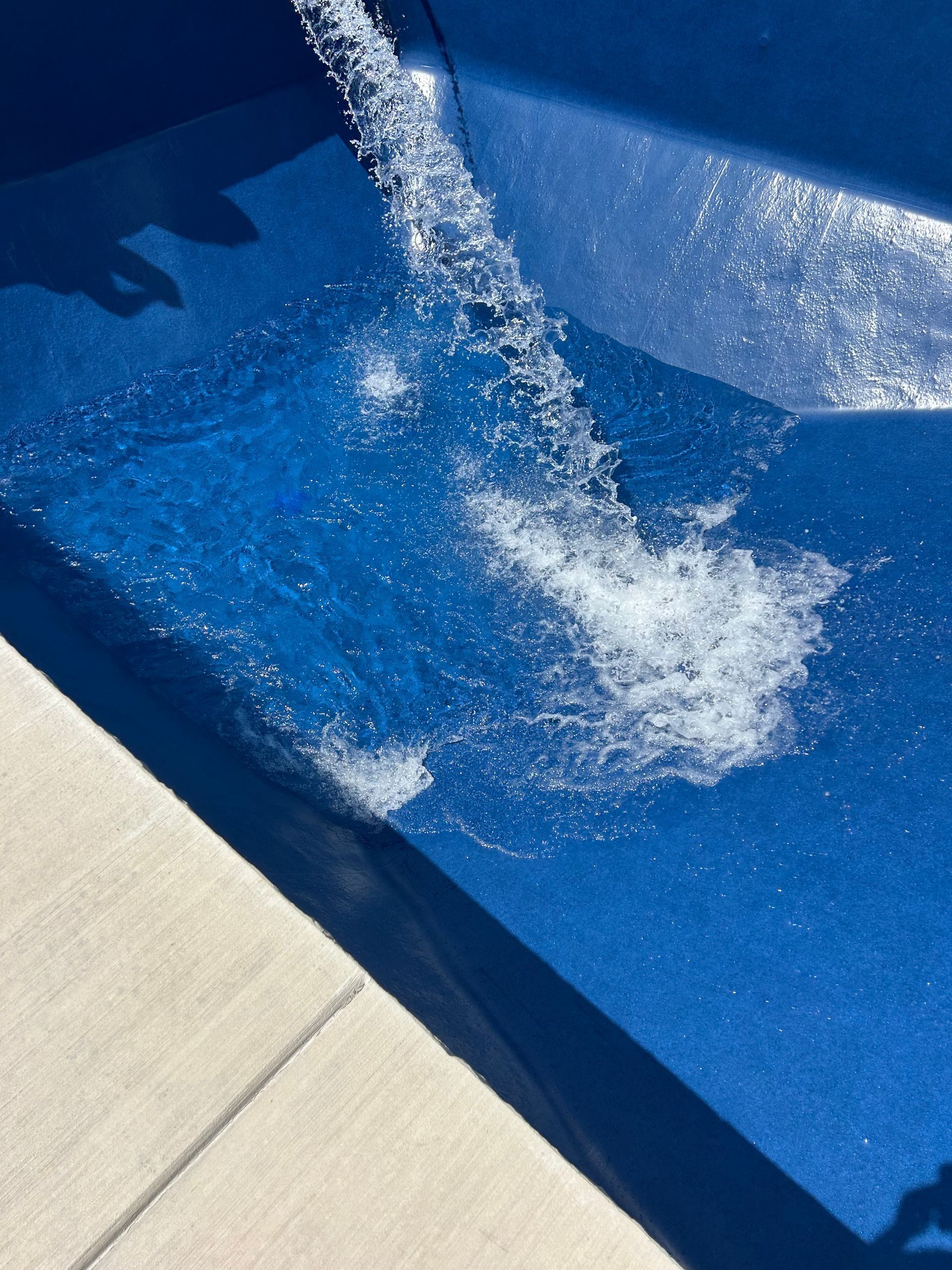 Water pouring into a blue-tiled pool, creating splashes and bubbles. Edge of concrete nearby.