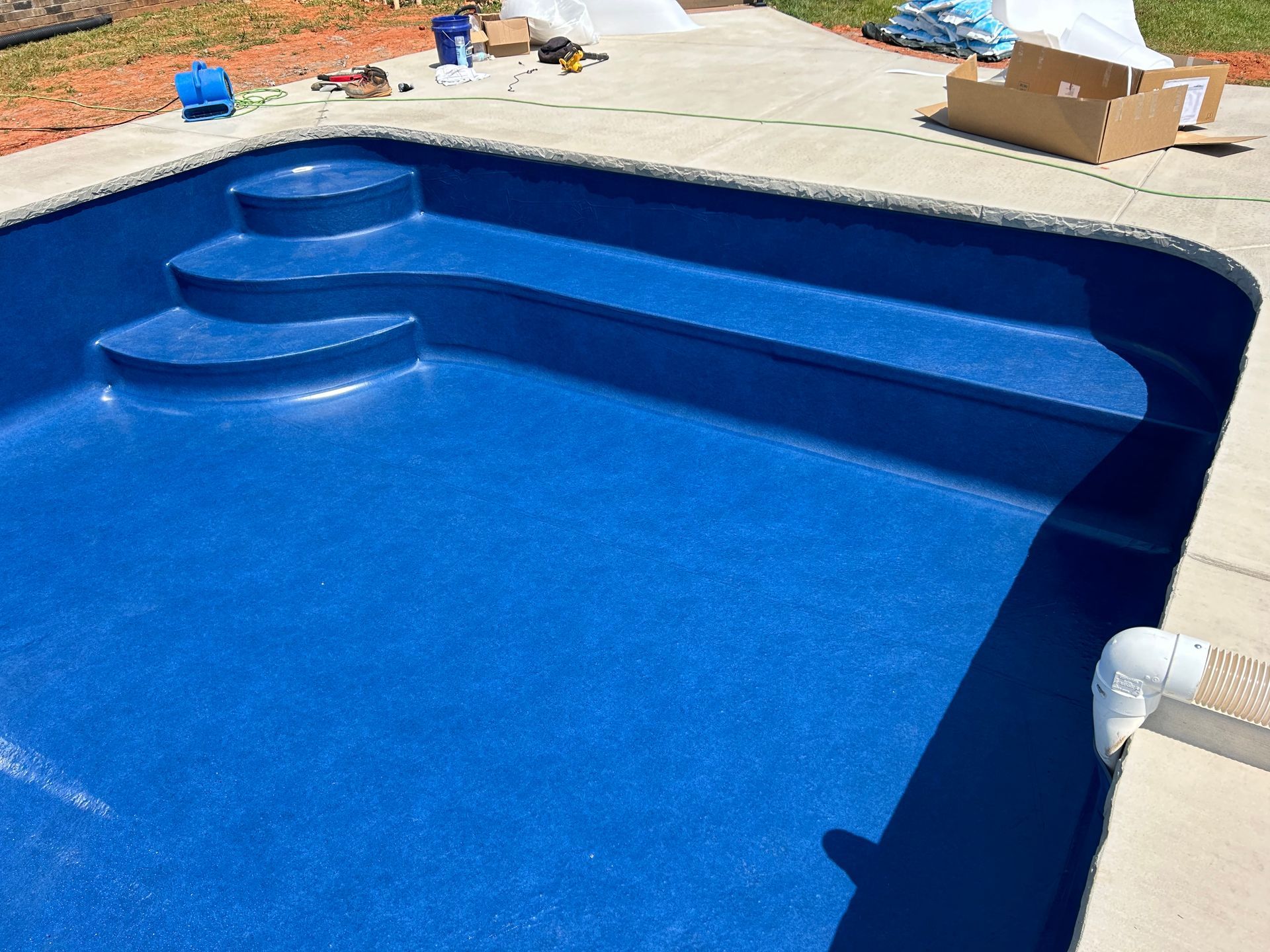 Blue swimming pool with steps, surrounded by concrete deck. Construction materials visible nearby.