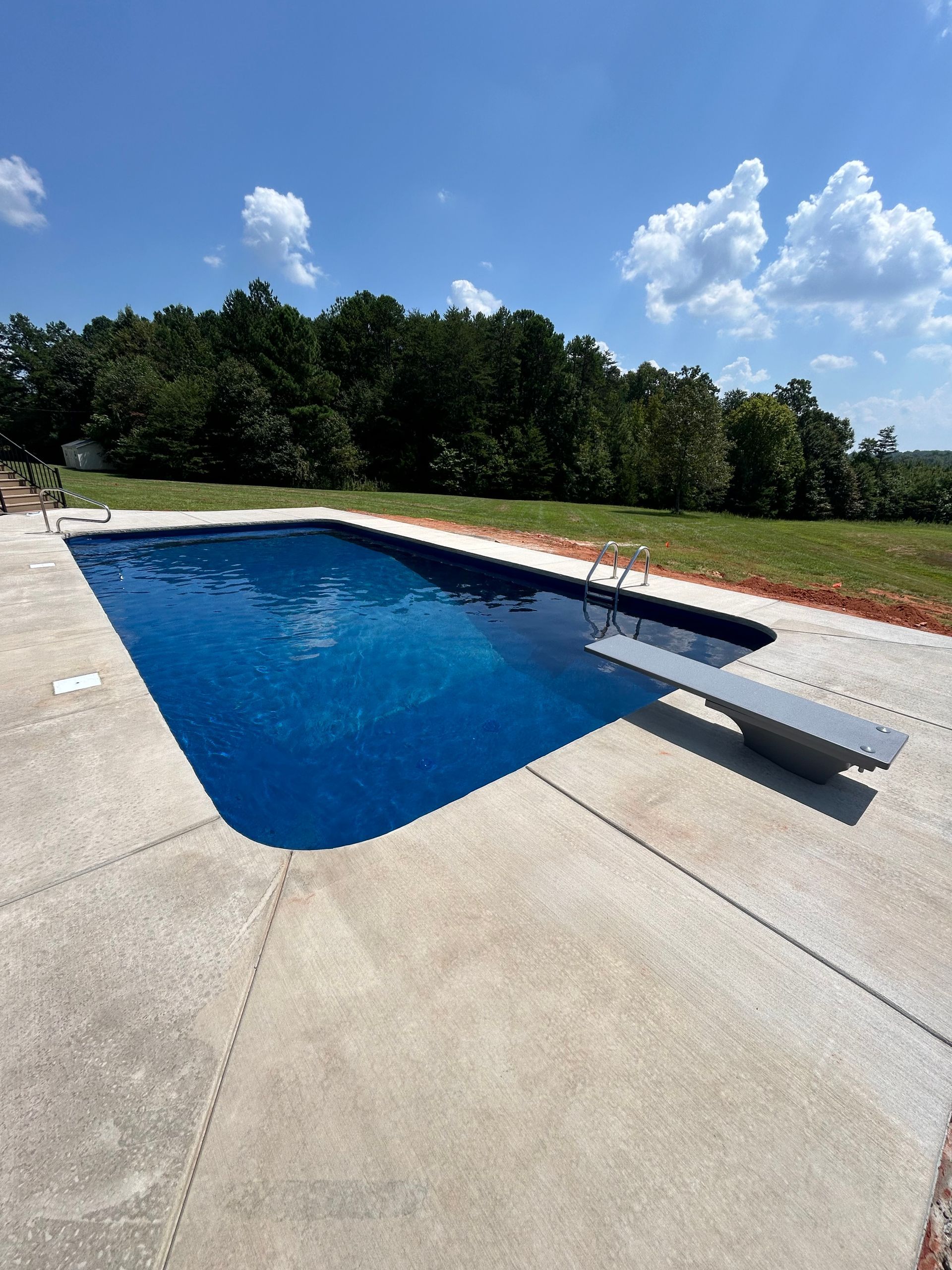 Rectangular swimming pool with diving board, blue water, concrete deck, and trees under a blue sky.