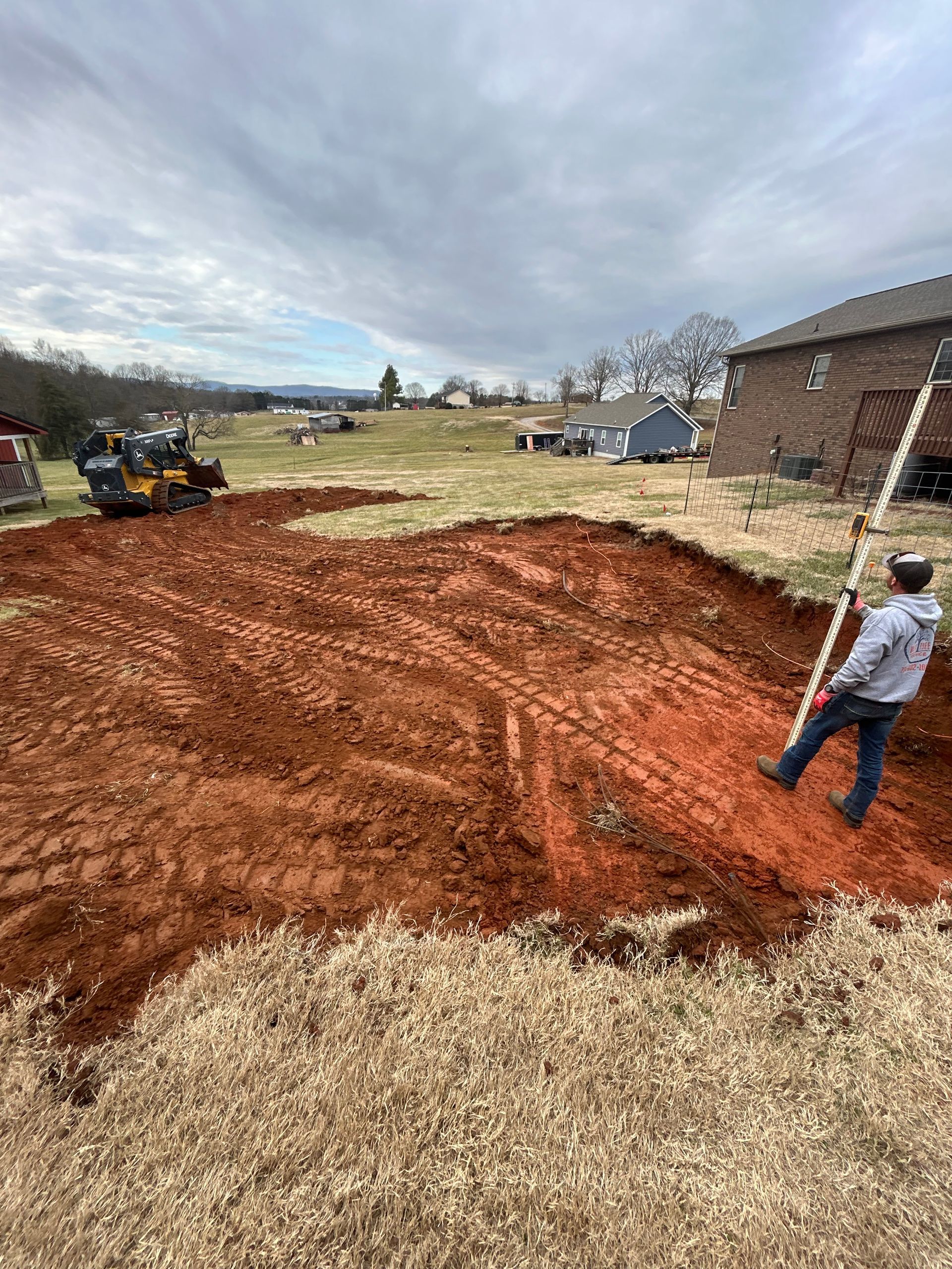 Construction site: person leveling red soil with a rod, excavator in background, cloudy sky.