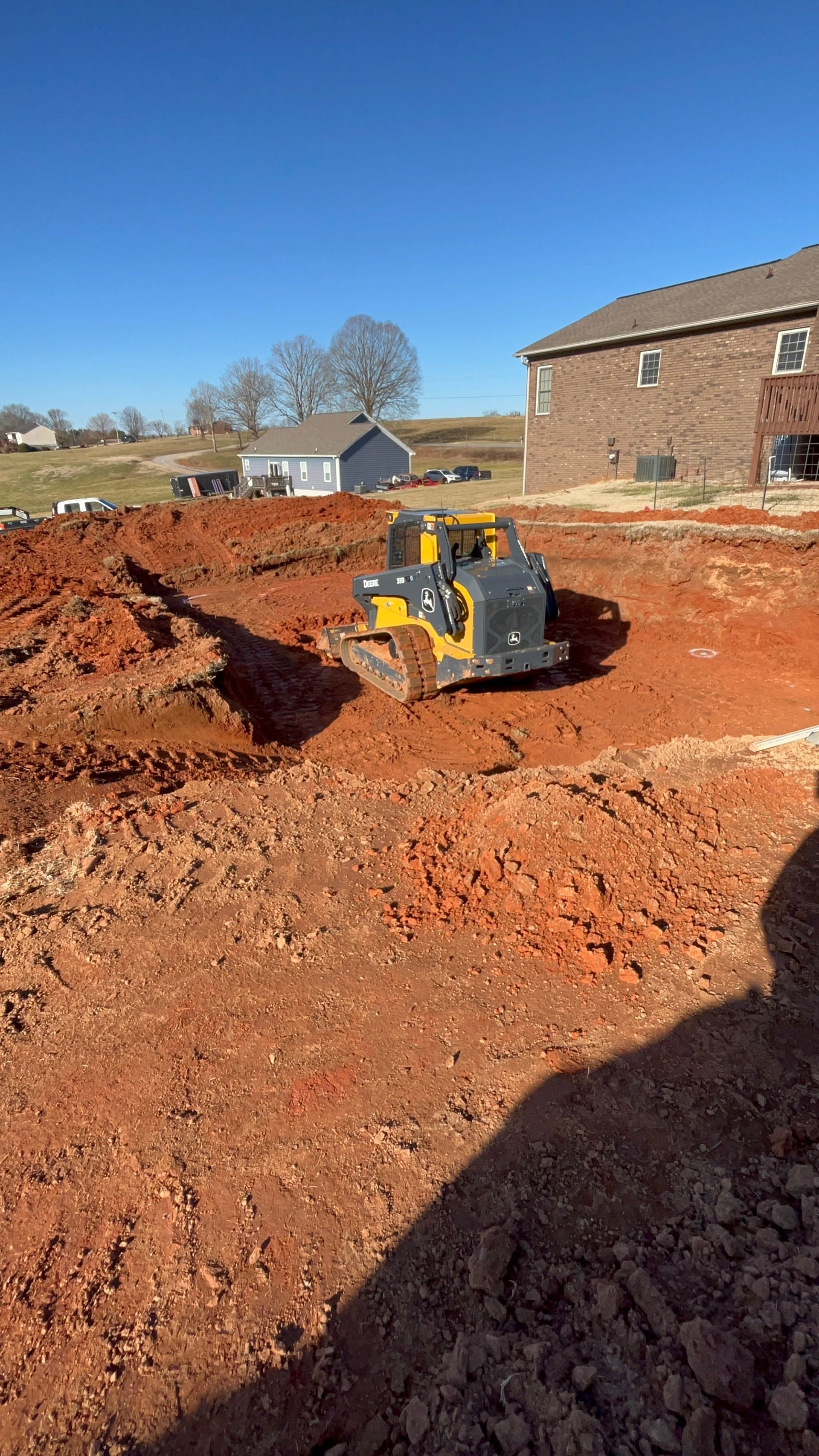 A yellow bulldozer on a construction site leveling red soil, residential buildings in the background.