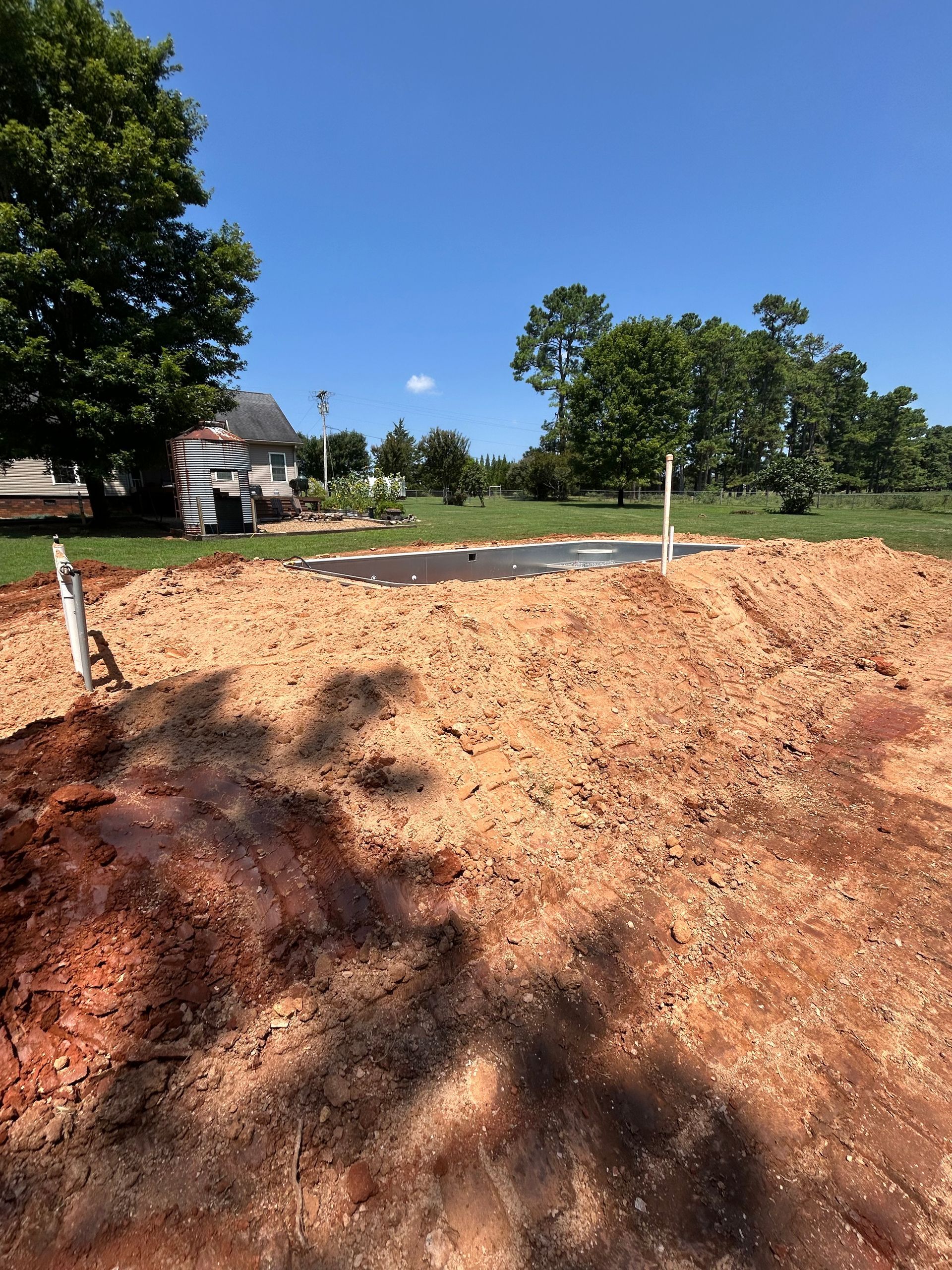 A cleared construction site with red dirt, plumbing, and a house in the background under a blue sky.