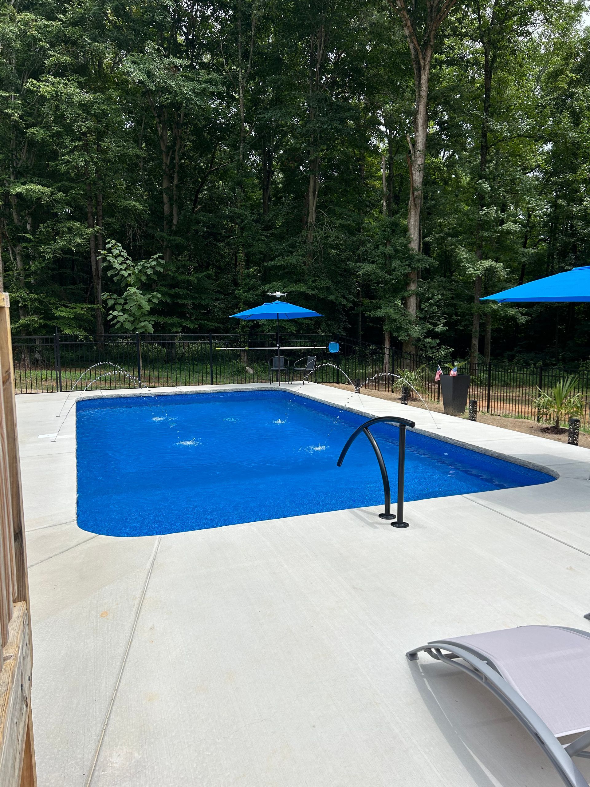 Blue-covered swimming pool surrounded by concrete patio, black fence, and trees. Two blue umbrellas.