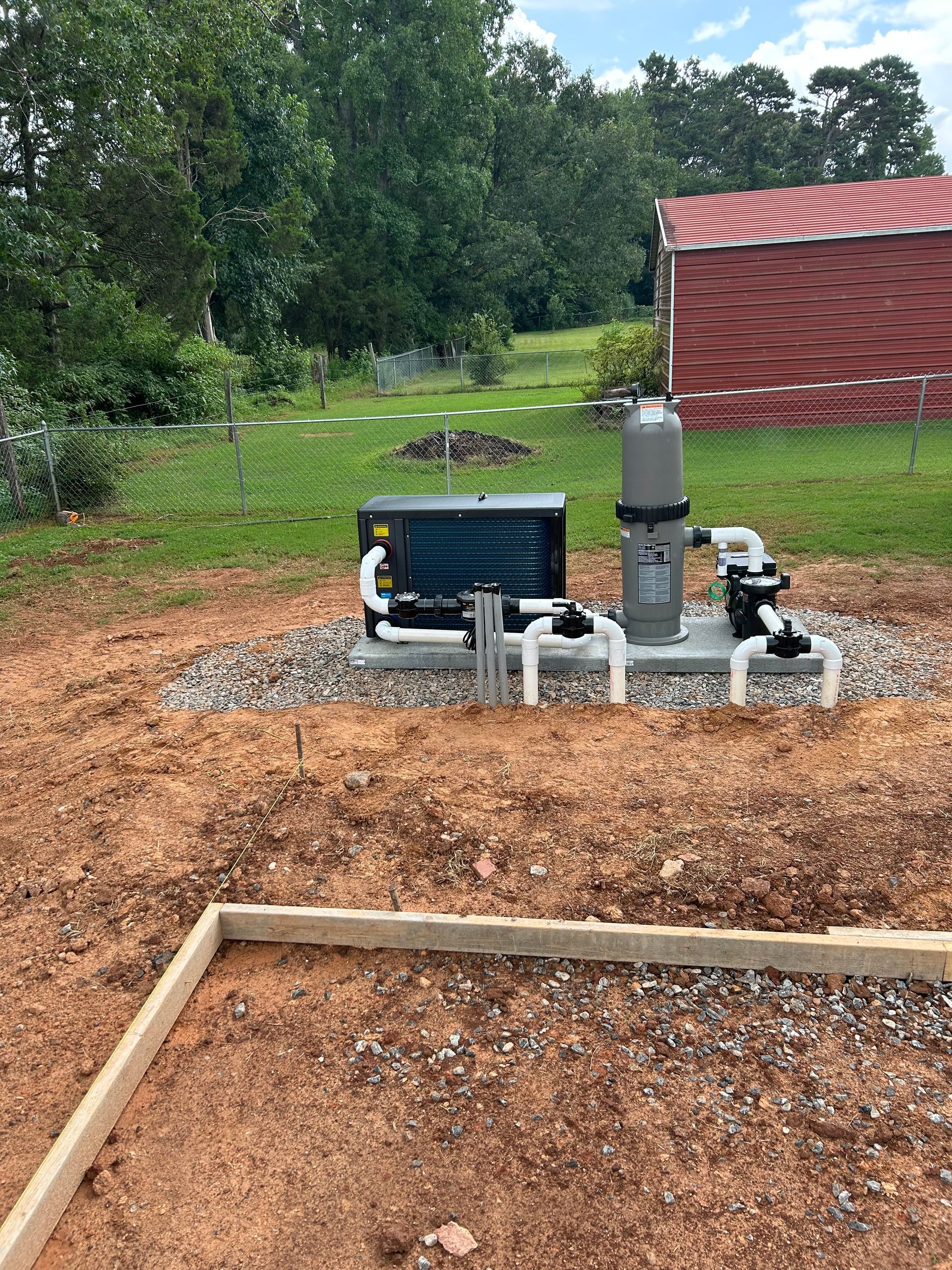 Pool equipment on gravel pad, including a filter, pump, and heater, outdoors near a red-roofed building.