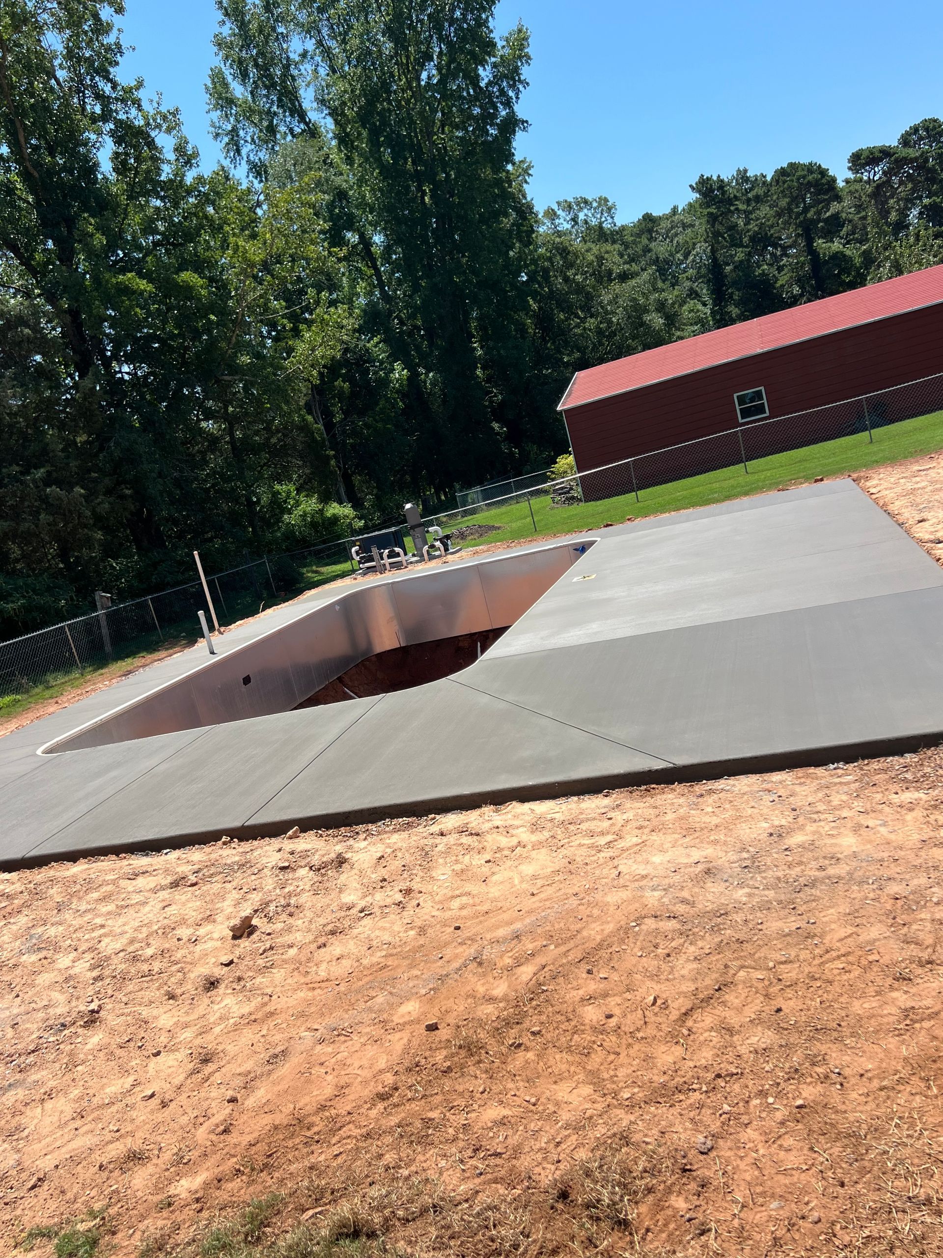 Concrete pool deck under construction with a red building in the background. Brown dirt surrounds.