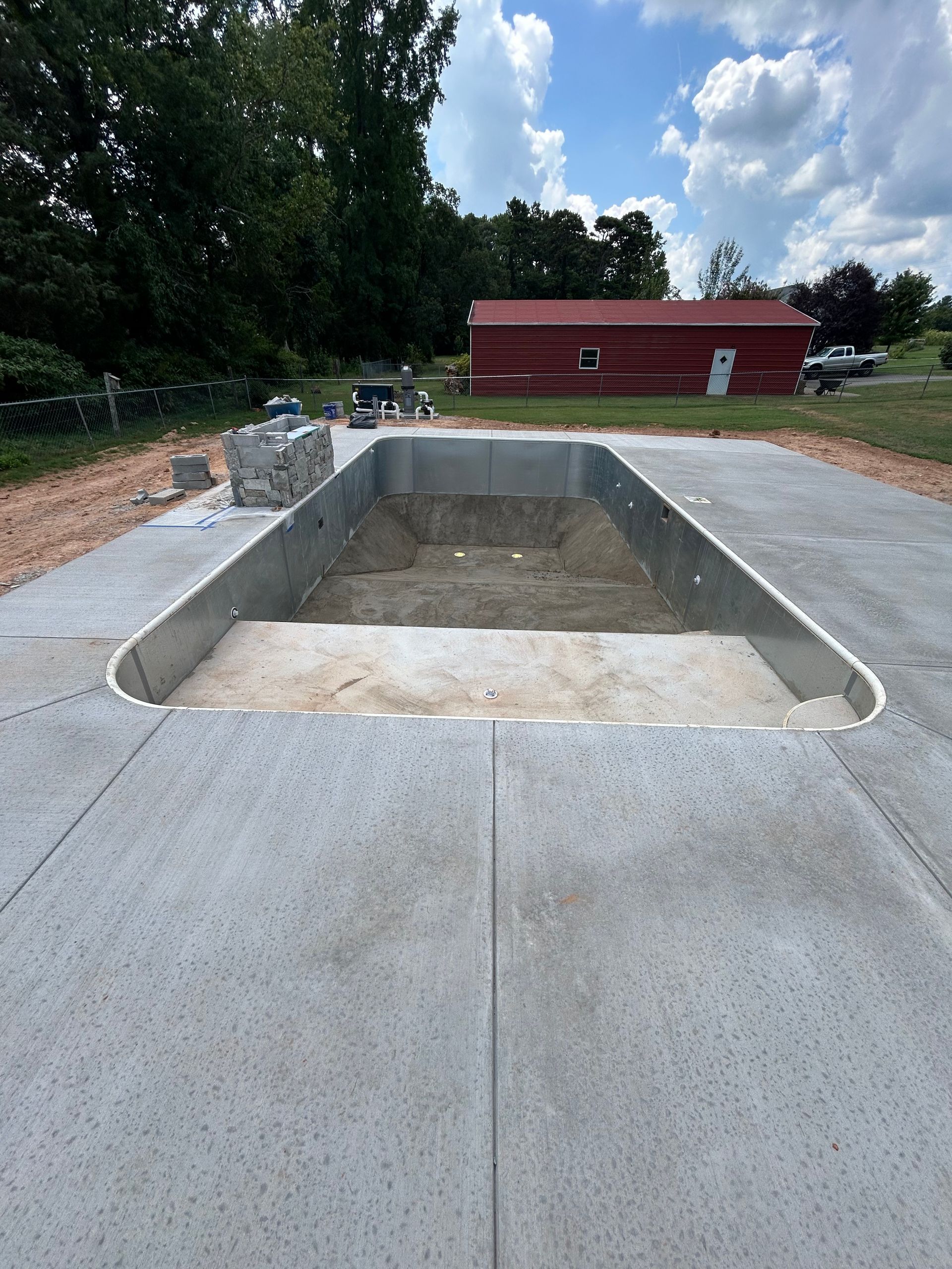 Empty concrete swimming pool under construction, with surrounding concrete patio. Red barn in background.