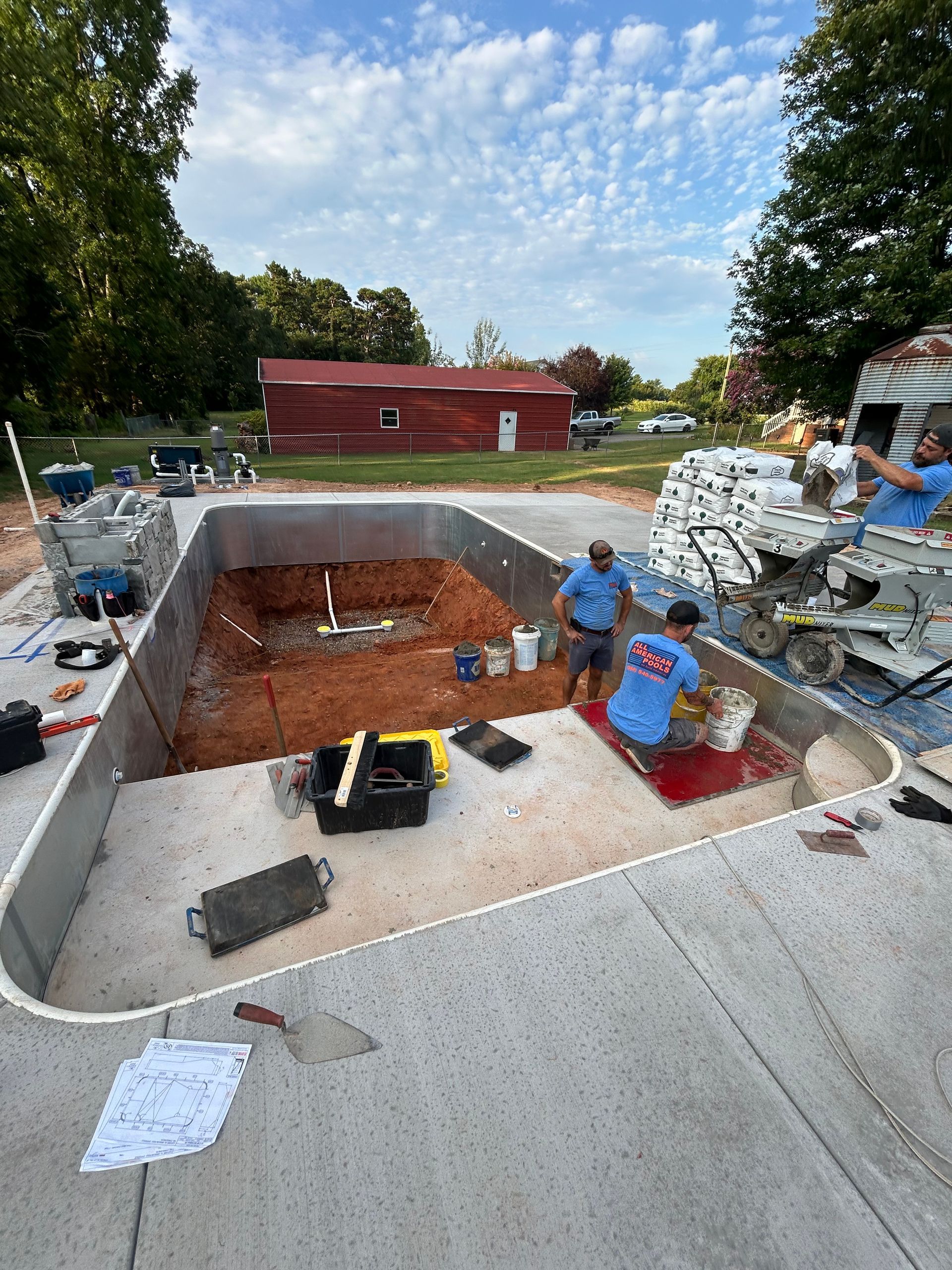 Pool construction site. Workers inside a rectangular pool shell, prepping and installing plumbing. Cement patio.