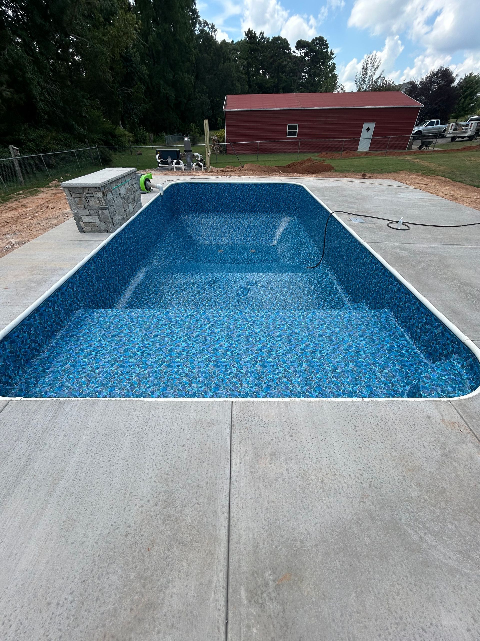 Rectangular pool with blue mosaic-like liner, concrete deck, and a red barn in the background.