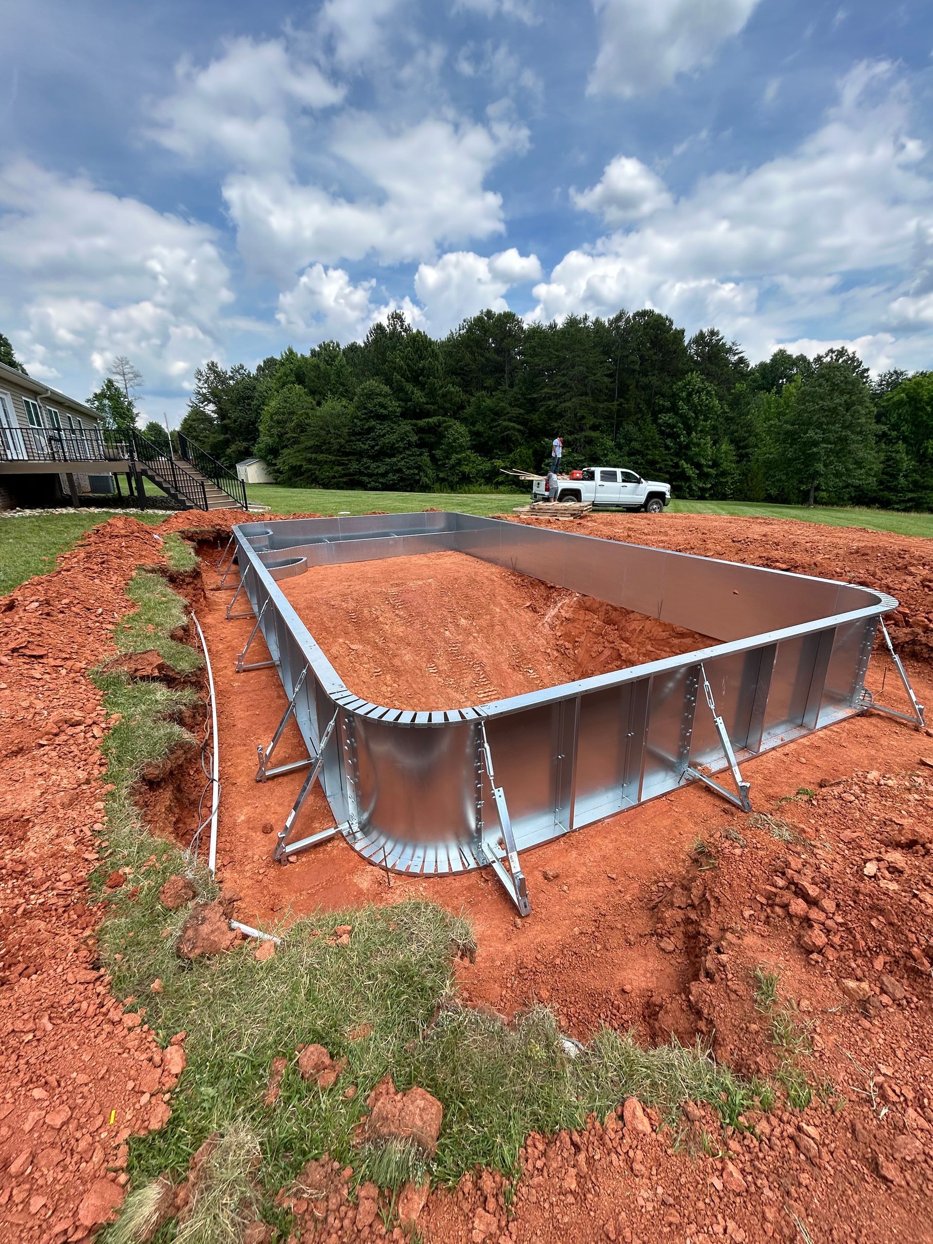 Pool frame installation in progress in a red dirt yard, with a truck and trees visible in the background.