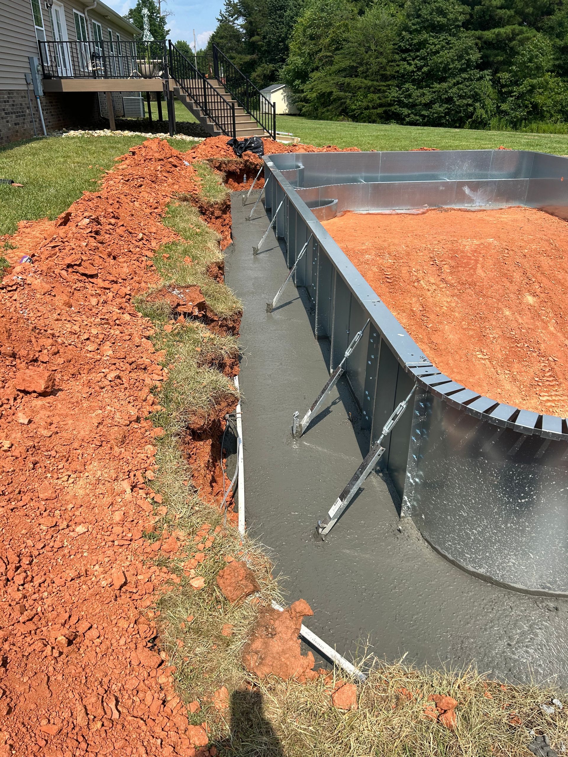 Above-ground pool under construction. Metal walls stand in a trench with new concrete. Red dirt surrounds.