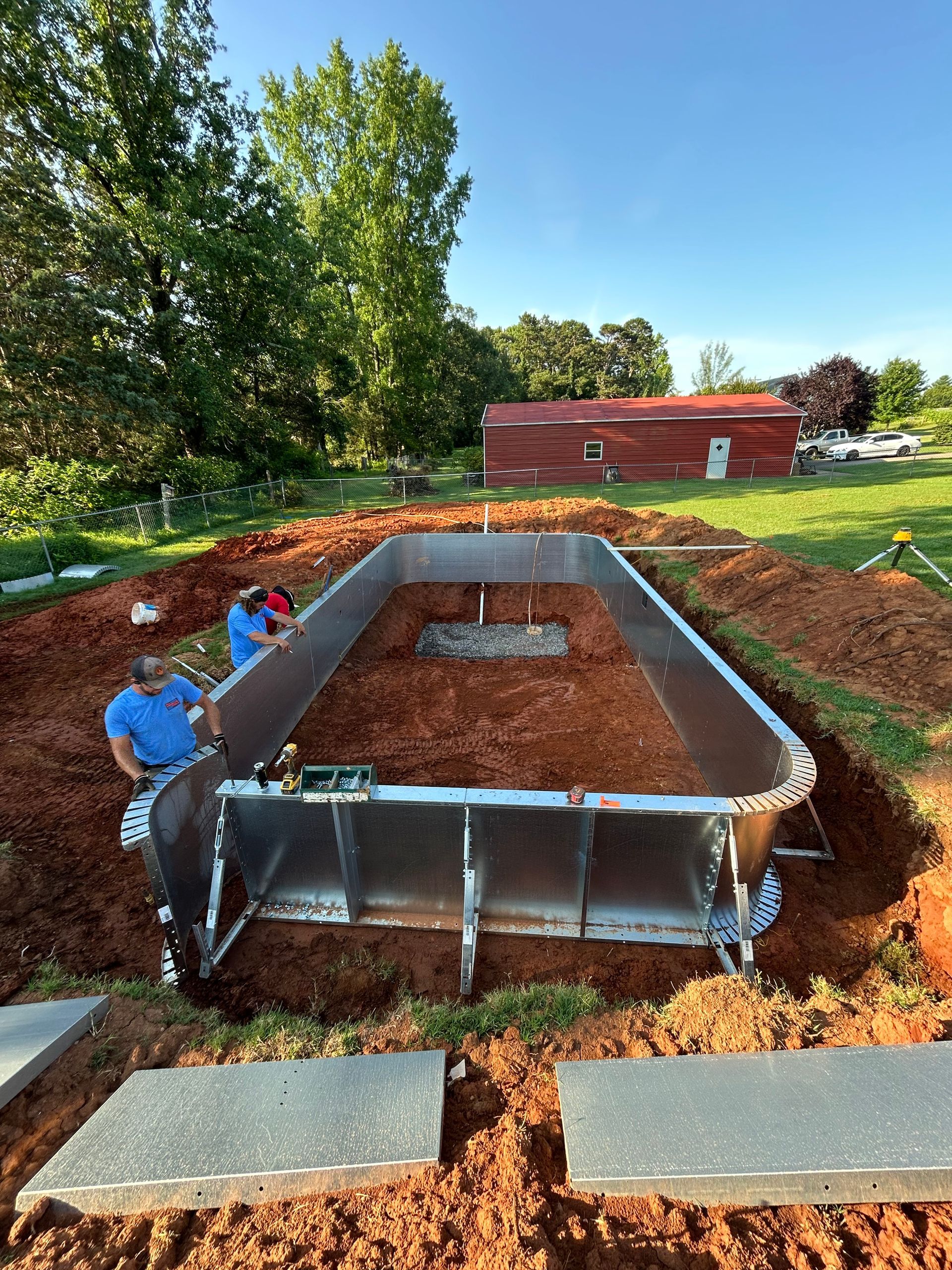 Workers installing a rectangular, above-ground pool; the frame is metal, surrounded by dirt, in a yard.