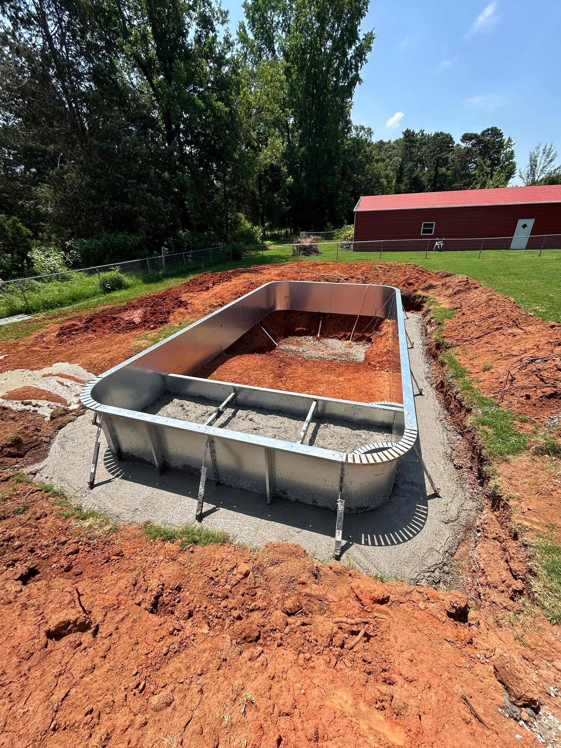 A rectangular metal structure set in a dirt excavation, likely a pool. A red barn is visible in the background.