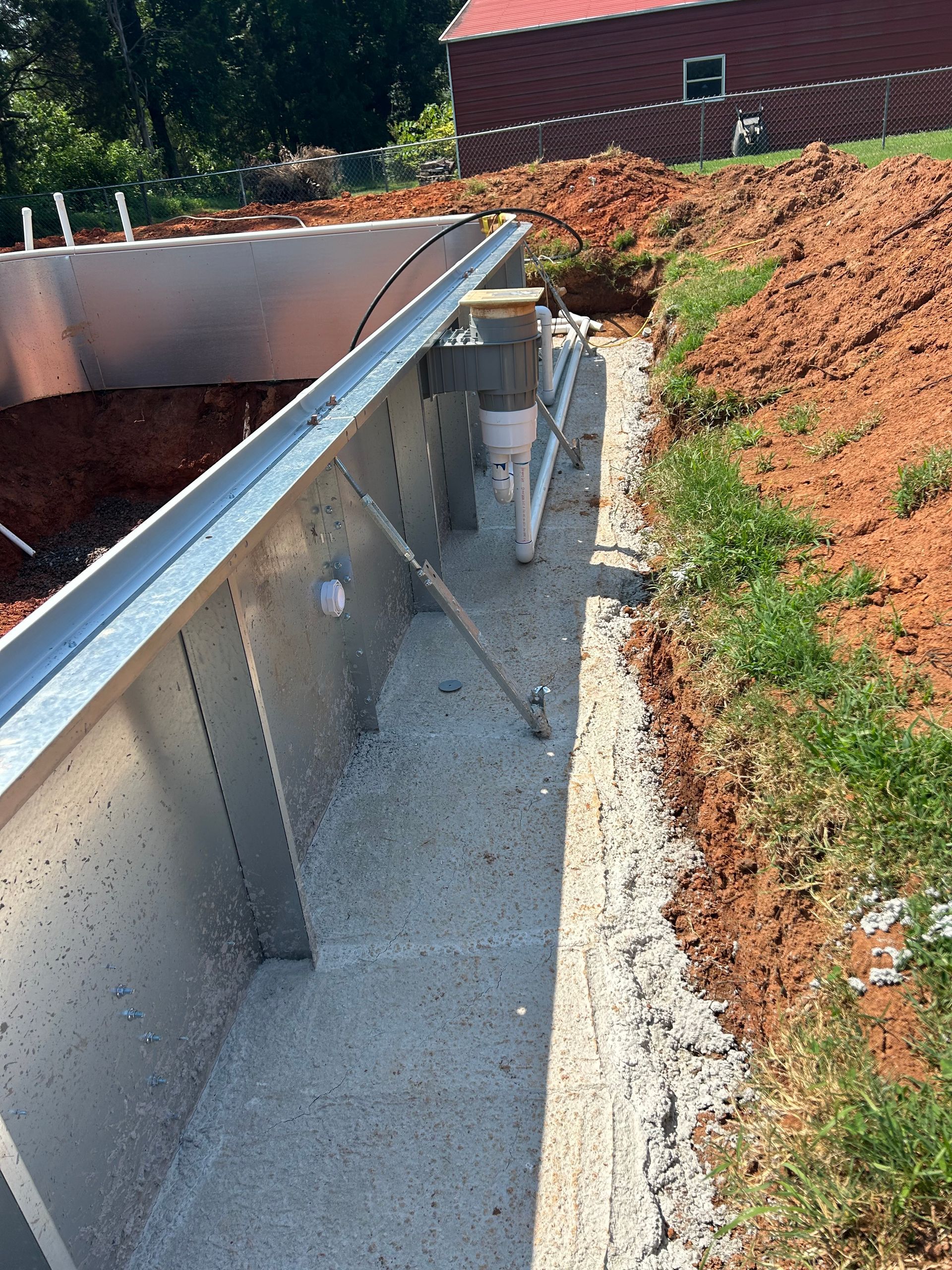 Metal pool structure under construction with plumbing and concrete base. Soil surrounds the structure.