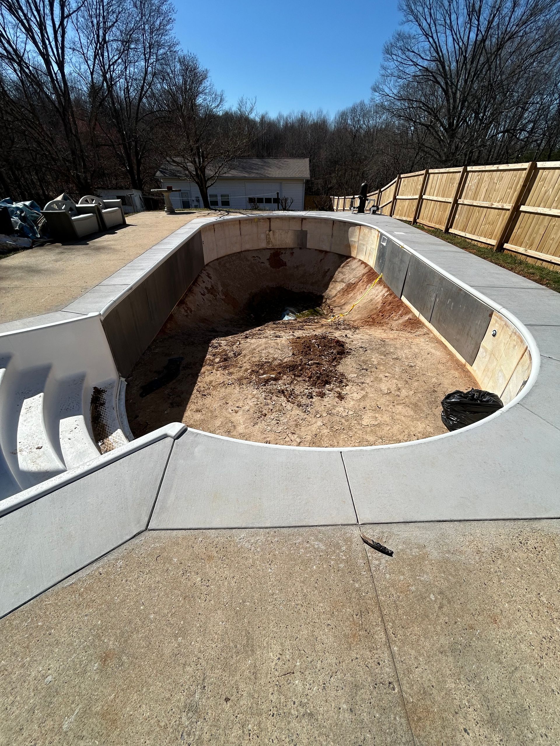 Empty, unkempt swimming pool with concrete surround. Adjacent fence and building in background. Sunny day.