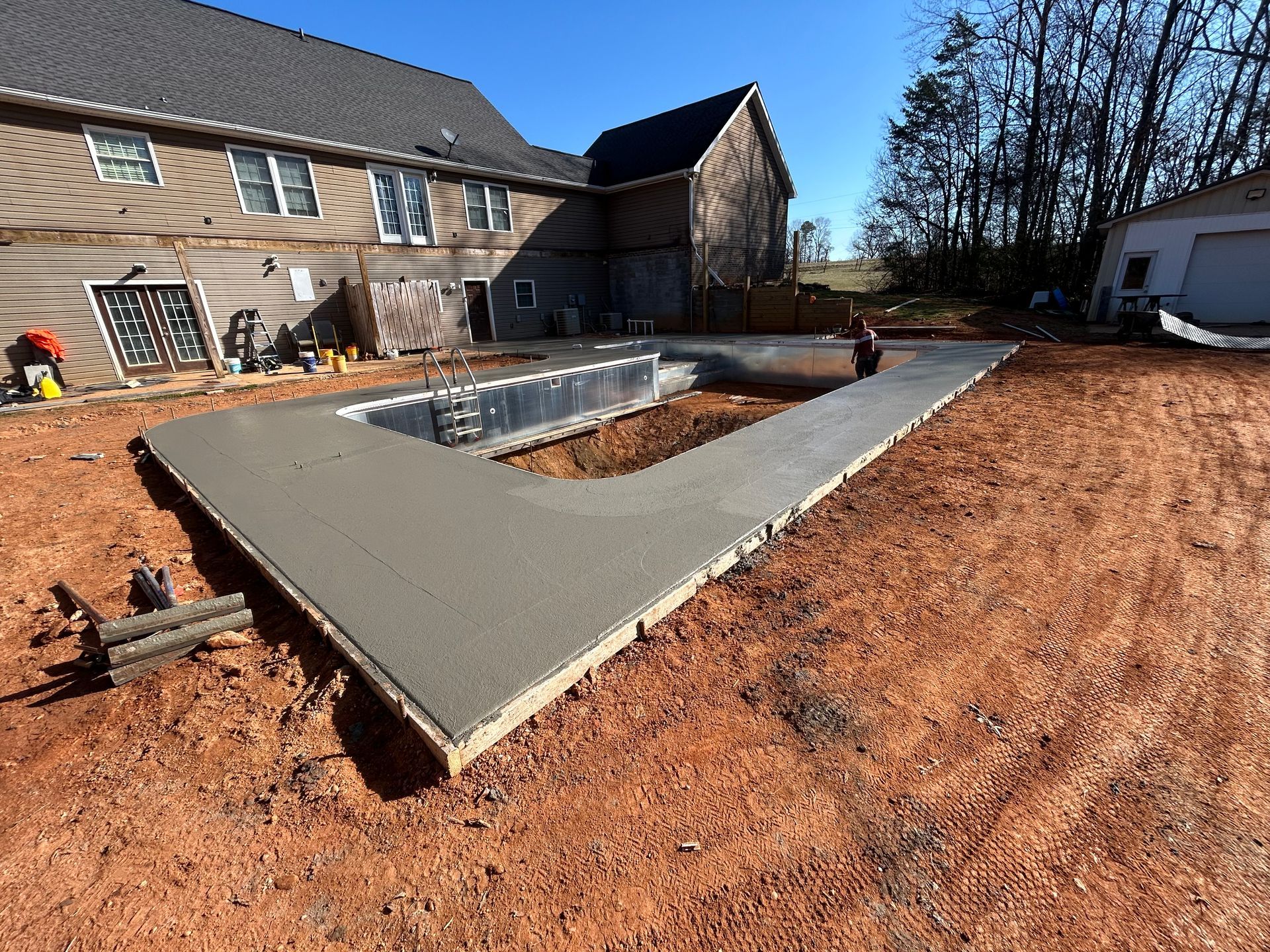 Concrete pool deck under construction next to a house and shed on a dirt lot.