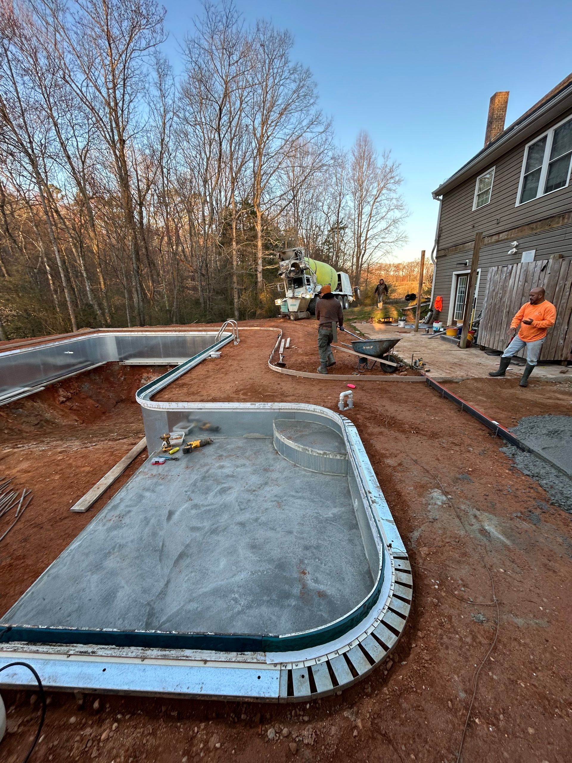 Construction of a concrete pool and surrounding patio. Workers are pouring concrete near a house, with trees in the background.